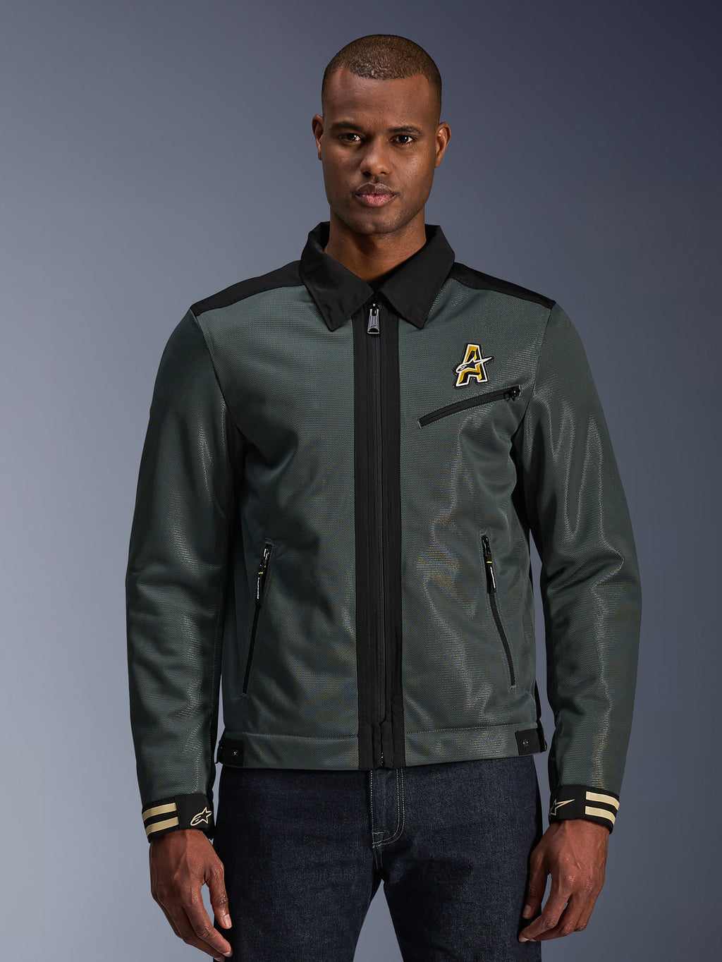 Alpinestars Ignite Air Jacket, giacca da moto urban, verde foresta scuro con dettagli neri, indossata da un modello con pantaloni in denim, colletto a camicia stile Harrington, zip frontale, logo sul petto e polsini a righe, moderna costruzione in mesh e tessuto per la guida estiva