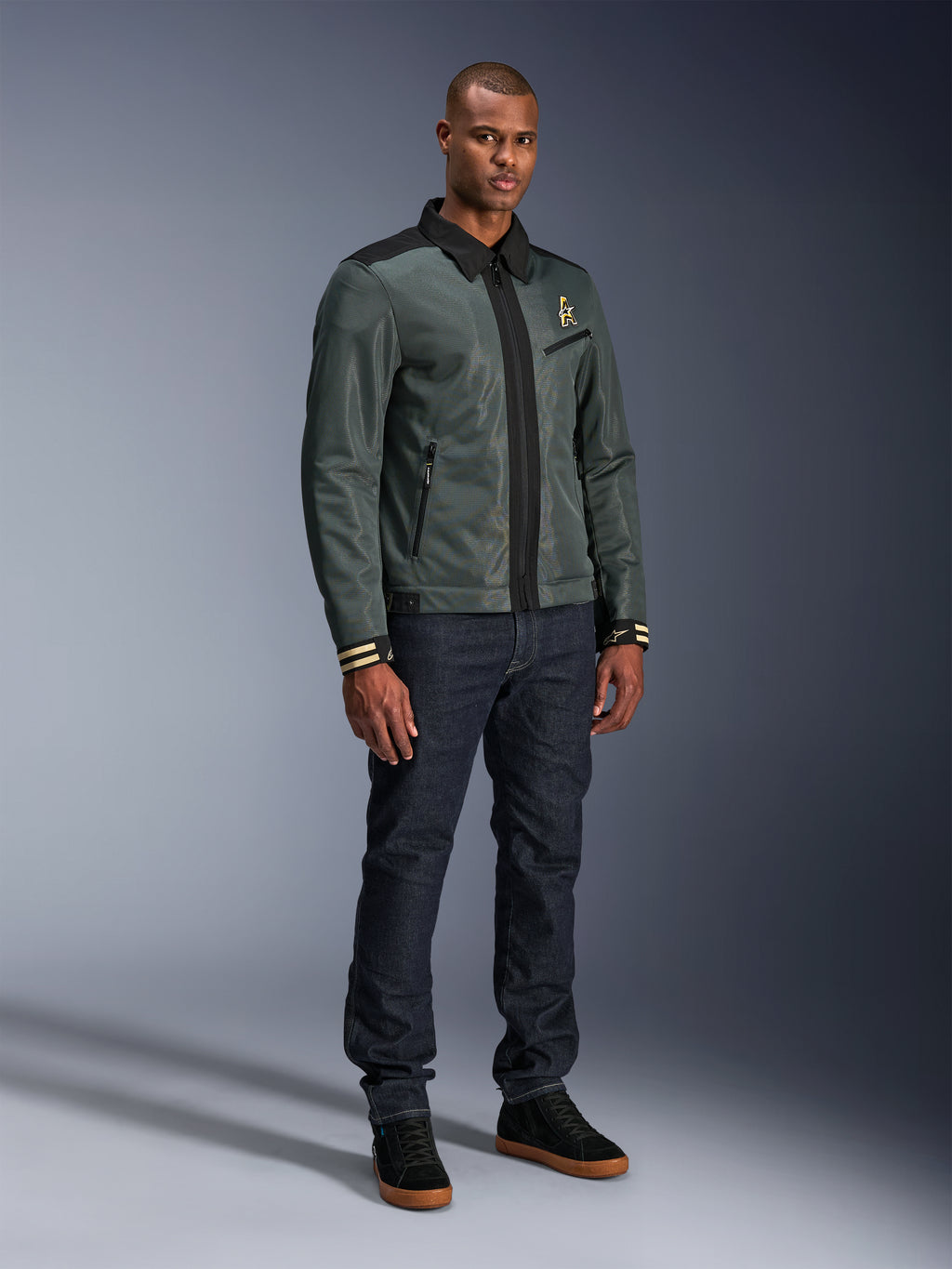 Giacca Alpinestars Ignite Air, Urban Gray Black Blue, giacca da moto urban, indossata da un modello con jeans in denim scuro e sneakers nere, caratterizzata da un corpo in rete, colletto a camicia tradizionale, silhouette d’ispirazione Harrington e logo Alpinestars dorato.