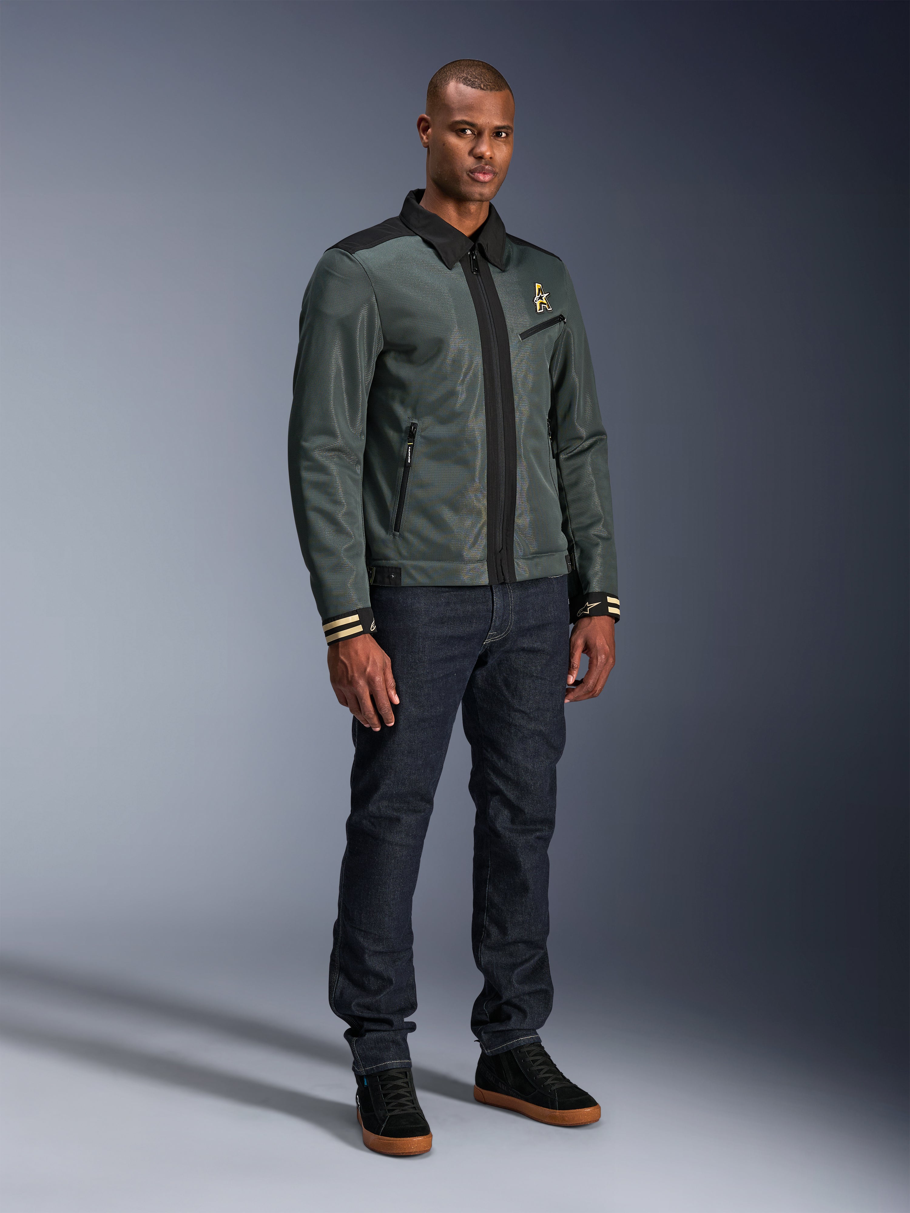 Giacca Alpinestars Ignite Air, Urban Gray Black Blue, giacca da moto urban, indossata da un modello con jeans in denim scuro e sneakers nere, caratterizzata da un corpo in rete, colletto a camicia tradizionale, silhouette d’ispirazione Harrington e logo Alpinestars dorato.