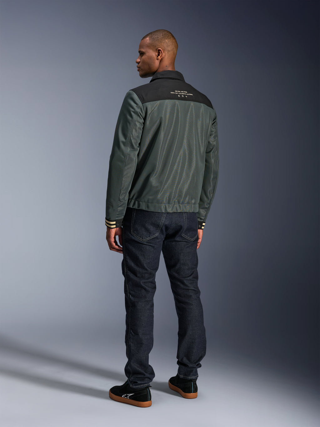 Giacca Alpinestars Ignite Air in Urban Gray Black Blue, vista posteriore su modello, con corpo in mesh, sprone nero sulle spalle e scritte bianche, polsini a righe, abbinata a jeans in denim scuro e sneakers nere, abbigliamento moto urban lifestyle