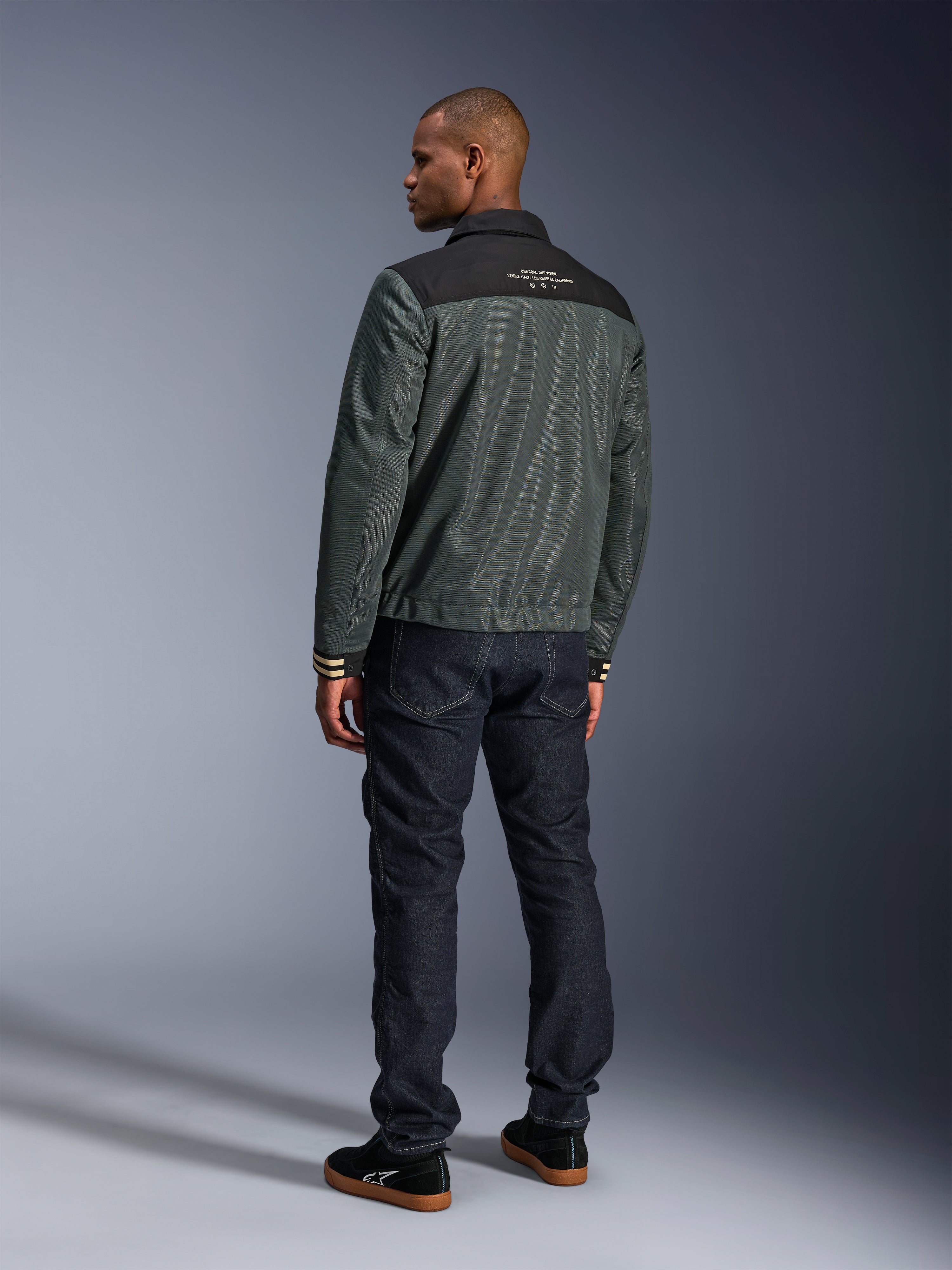Giacca Alpinestars Ignite Air in Urban Gray Black Blue, vista posteriore su modello, con corpo in mesh, sprone nero sulle spalle e scritte bianche, polsini a righe, abbinata a jeans in denim scuro e sneakers nere, abbigliamento moto urban lifestyle