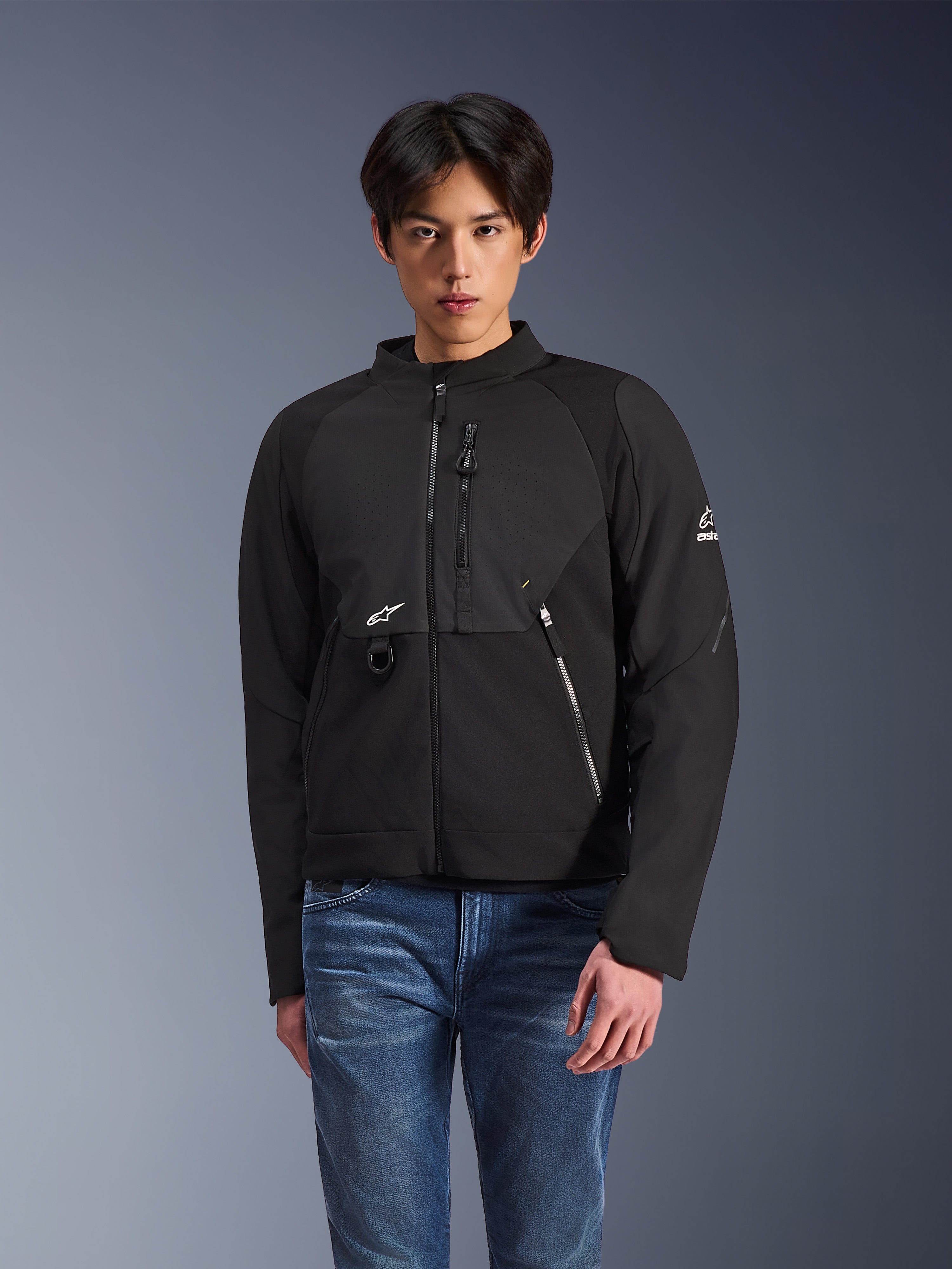Giacca Alpinestars Provoke, giacca moto urbana in tessuto nero, indossata da un modello con jeans blu, con pannelli sul petto perforati al laser, tasche multiple con zip, logo A-star bianco su petto e manica, design moderno per la guida su strada