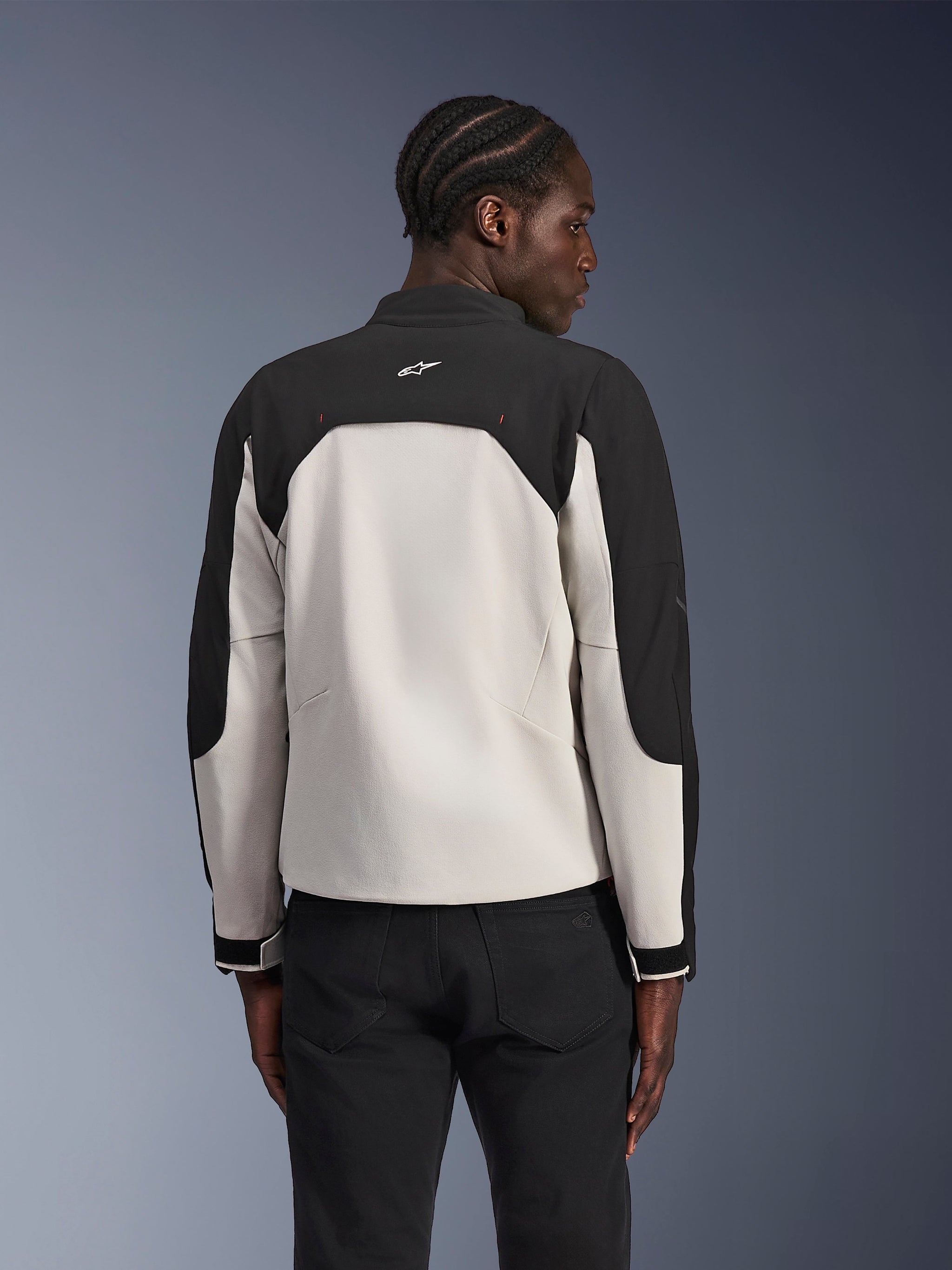 Giacca Alpinestars Provoke, giacca da moto urban, Black Metal Gray, vista posteriore su un modello, design in tessuto bicolore con spalle nere e pannello posteriore grigio chiaro, logo a stella Alpinestars sul colletto, abbinata a pantaloni neri su sfondo sfumato blu