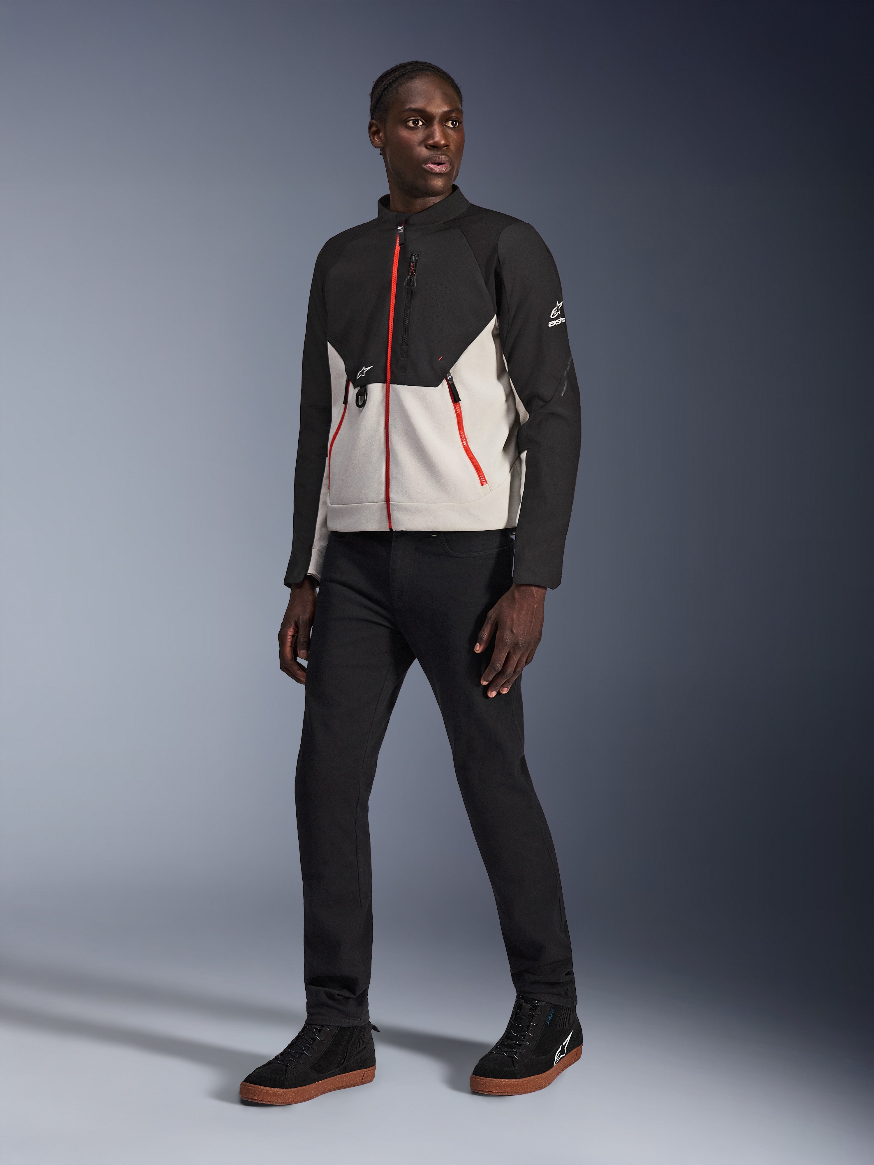 Modello che indossa la giacca Alpinestars Provoke in Black Metal Gray, caratterizzata da distintive cerniere rosse frontali e sulle tasche, abbinata a pantaloni neri e sneakers alte nere con suola color para, abbigliamento da moto urban contemporary per un look street-ready.