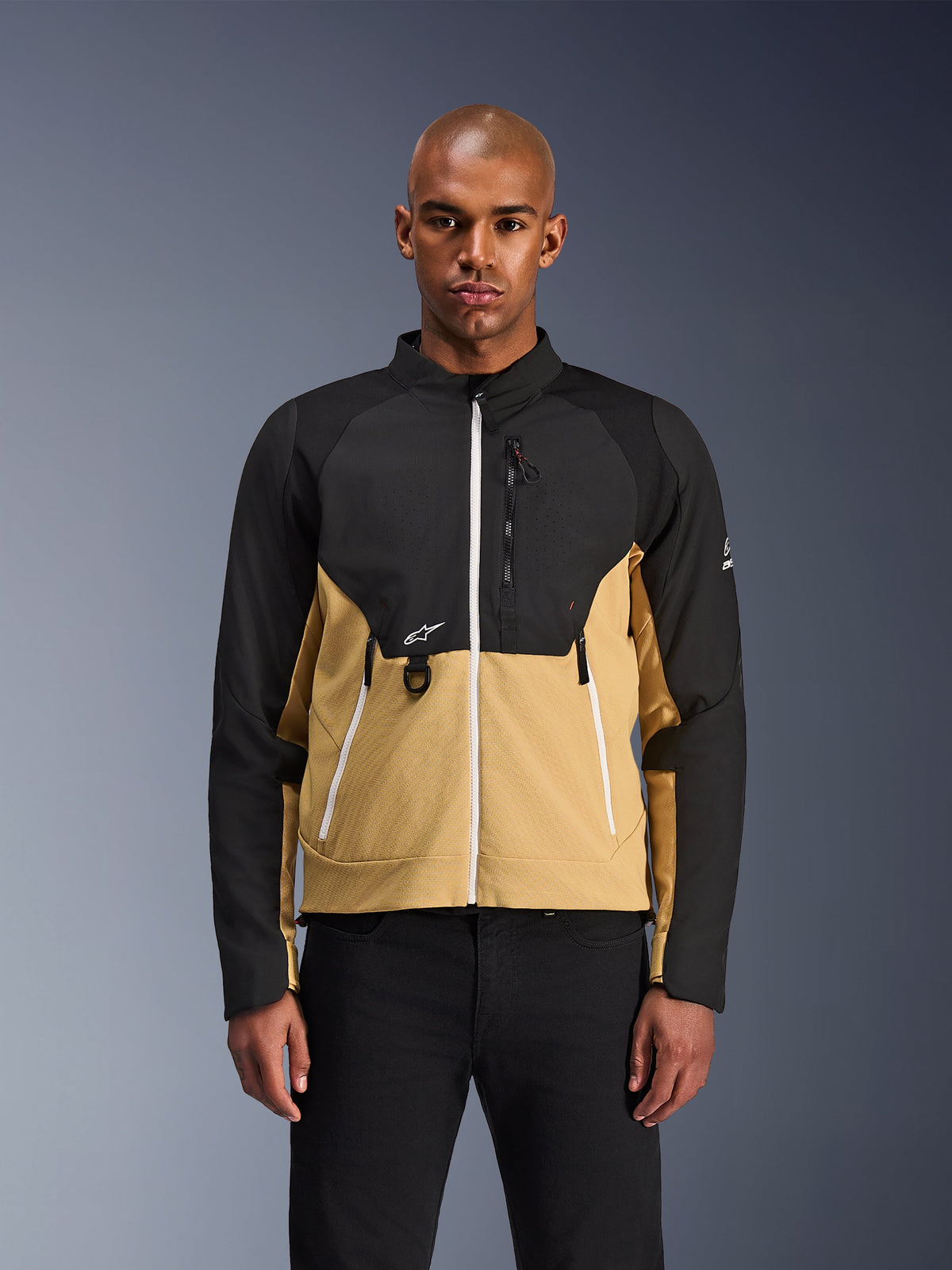 Alpinestars Giacca Provoke, giacca da moto urbana, nero e color cammello, vista frontale su un modello maschile, design bicolore con cerniere bianche, tasca sul petto con zip e anello a D e piccolo logo Alpinestars bianco, abbigliamento per motociclista urbano contemporaneo