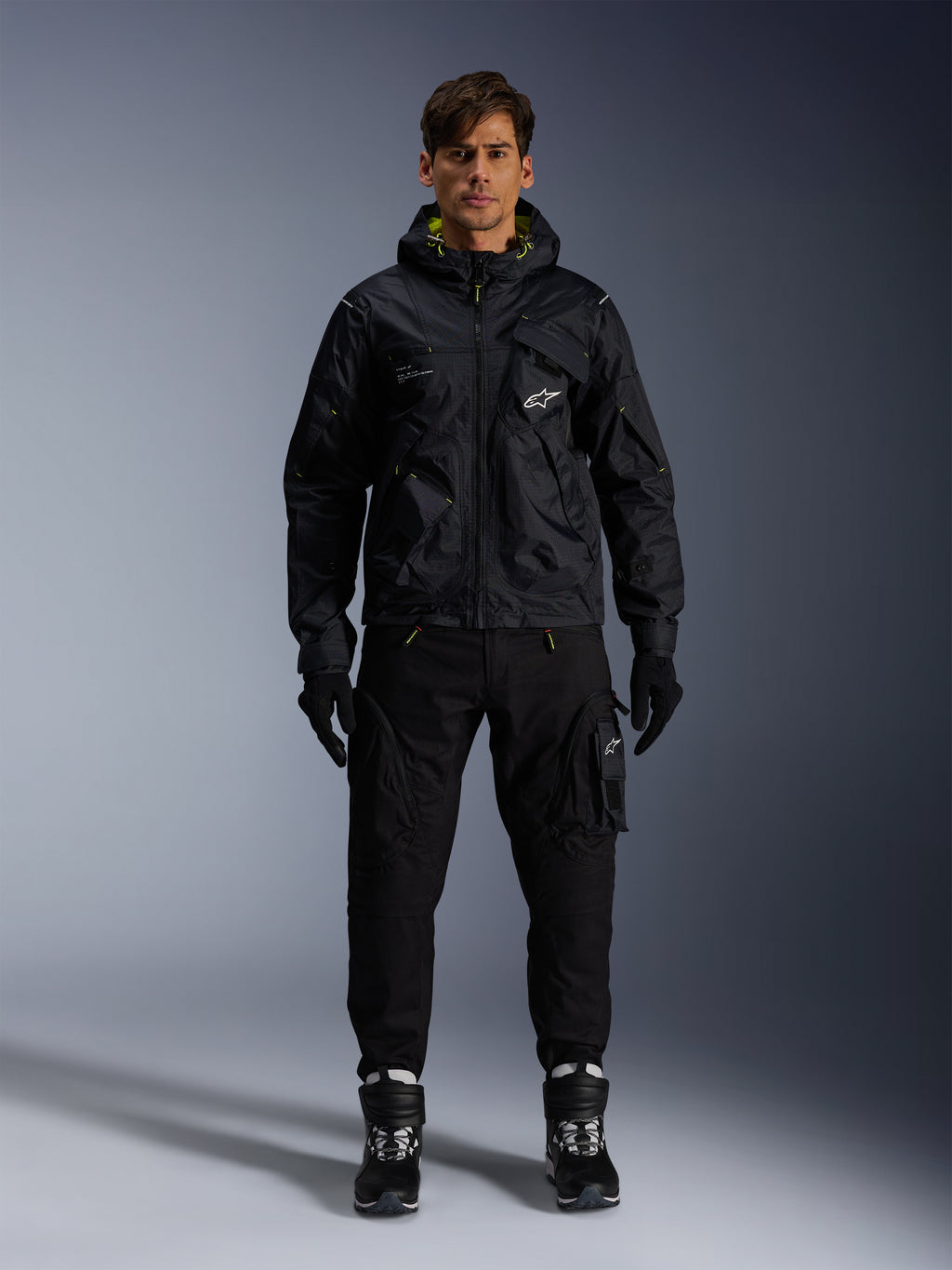 Modello che indossa la Giacca Alpinestars Mogress Air, giacca in tessuto nero con cappuccio e dettagli lime punch, abbinata a pantaloni utility neri, guanti neri e stivali da moto neri e bianchi, design streetwear ispirato al motorsport urbano