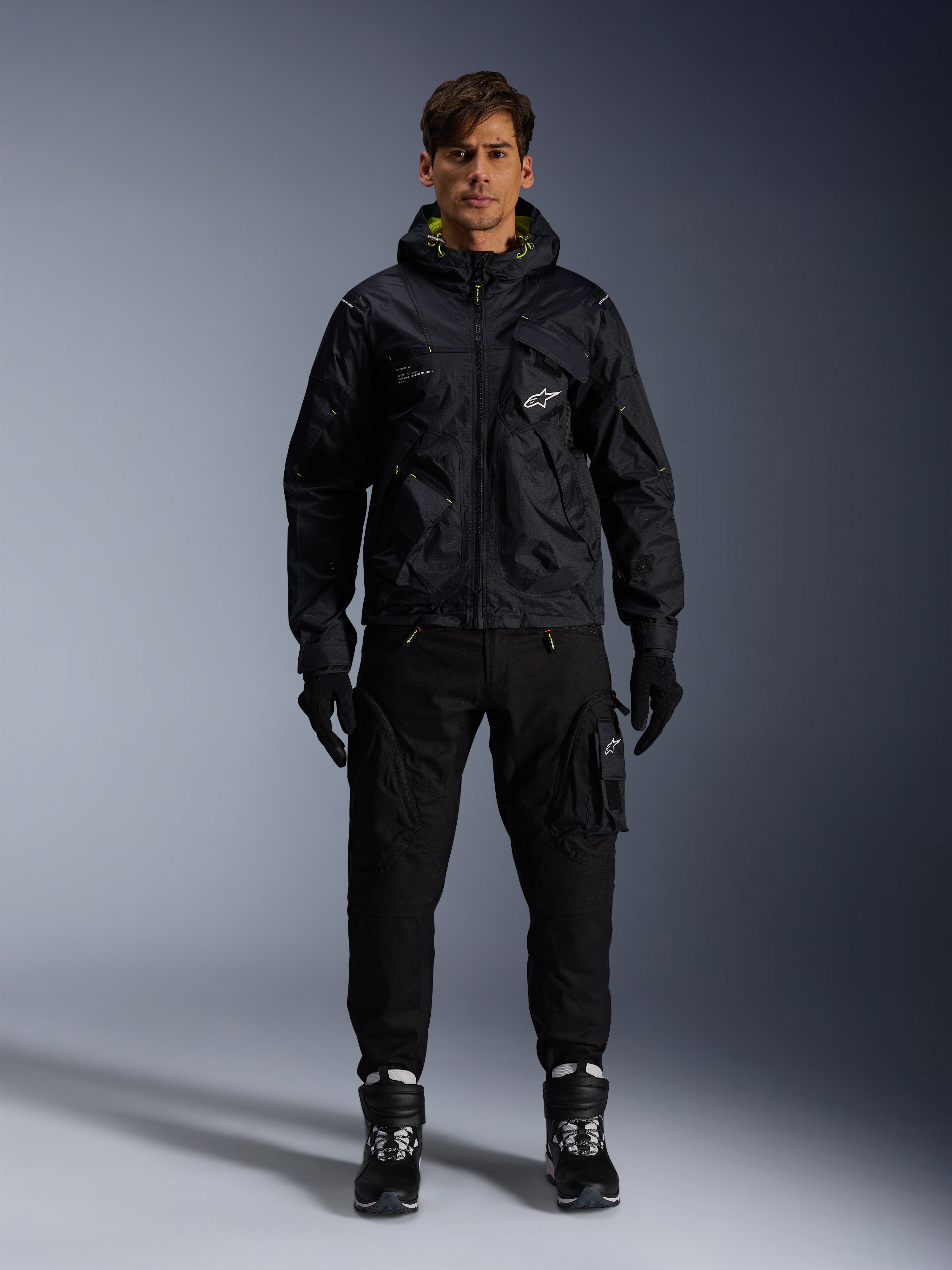 Modello che indossa la Giacca Alpinestars Mogress Air, giacca in tessuto nero con cappuccio e dettagli lime punch, abbinata a pantaloni utility neri, guanti neri e stivali da moto neri e bianchi, design streetwear ispirato al motorsport urbano