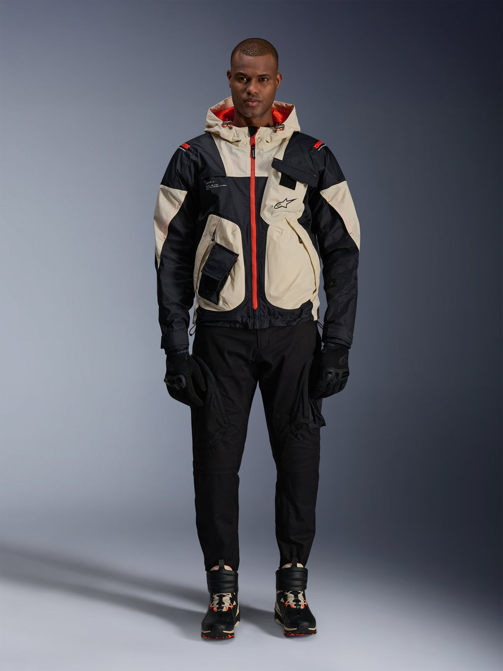 Giacca Alpinestars Mogress Air, giacca da moto urban, color Black Almond e Hot Orange, indossata da un modello con pantaloni e guanti neri, design d'ispirazione utility con pannelli crema e cerniere arancione acceso, streetwear moderno influenzato dal motorsport.