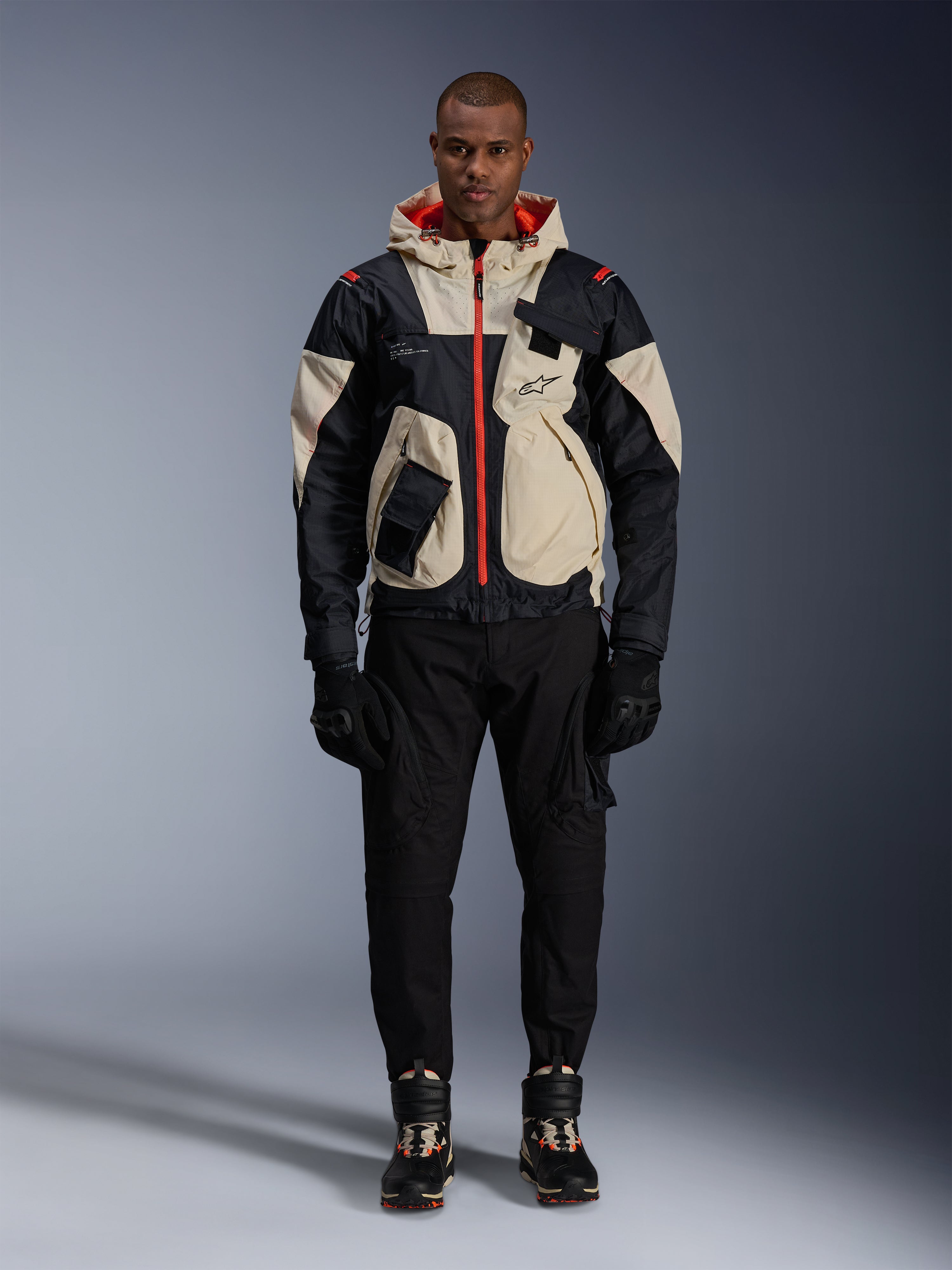 Giacca Alpinestars Mogress Air, giacca da moto urban, color Black Almond e Hot Orange, indossata da un modello con pantaloni e guanti neri, design d'ispirazione utility con pannelli crema e cerniere arancione acceso, streetwear moderno influenzato dal motorsport.
