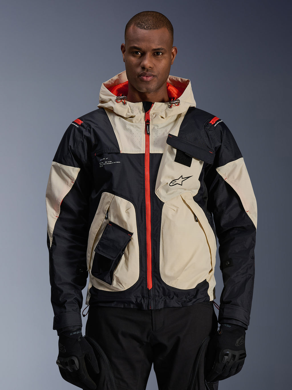 Alpinestars Mogress Air Jacket, giacca da moto urbana, color mandorla e nero con cerniere e fodera del cappuccio arancione acceso, design con cappuccio e tasche utility, logo sul petto, indossata con guanti neri