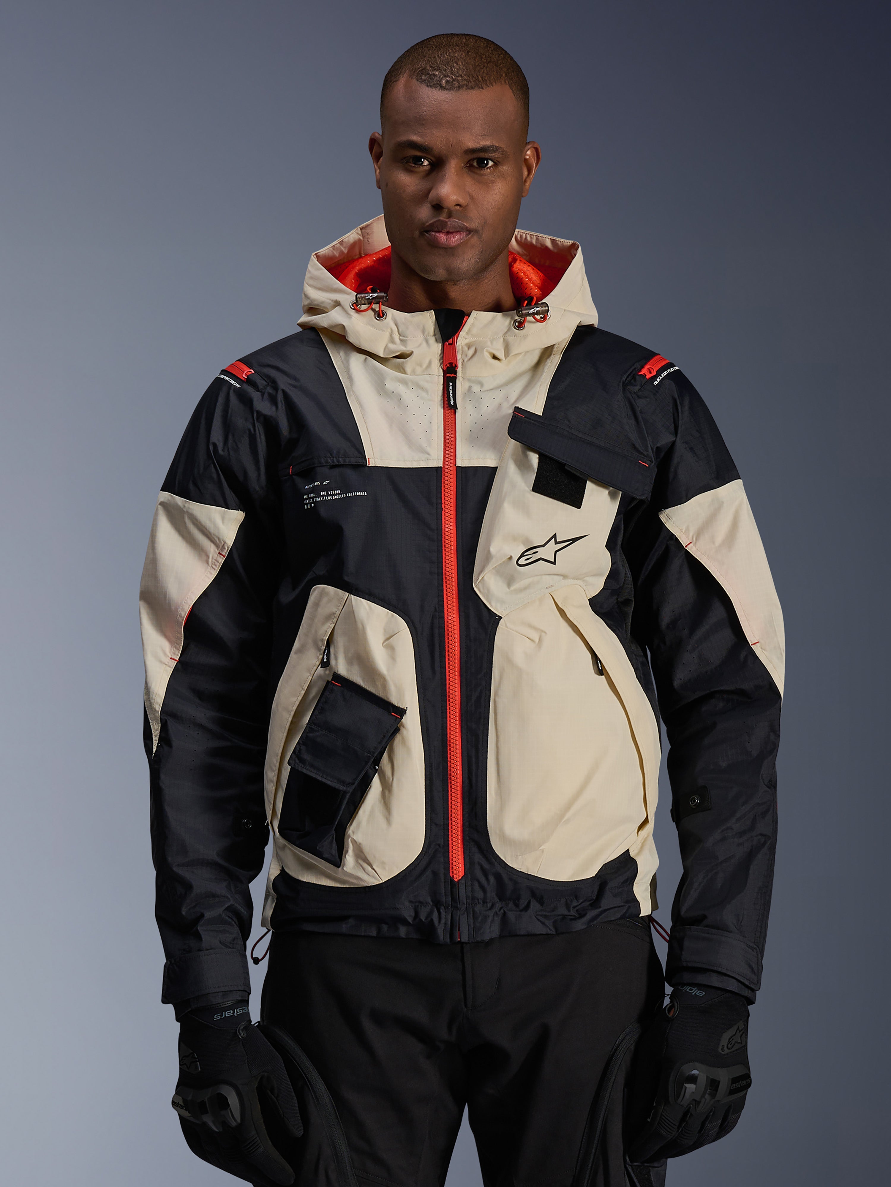 Alpinestars Mogress Air Jacket, giacca da moto urbana, color mandorla e nero con cerniere e fodera del cappuccio arancione acceso, design con cappuccio e tasche utility, logo sul petto, indossata con guanti neri