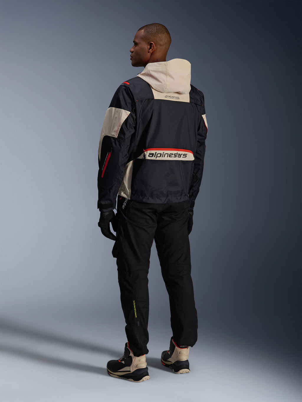 Alpinestars Mogress Air Jacket, Black Almond Hot Orange, vista posteriore del motociclista che indossa abbigliamento tecnico urbano, cappuccio color mandorla e pannello posteriore con grande logo Alpinestars, dettagli zip arancioni, abbinata a pantaloni neri e stivali da moto alti.