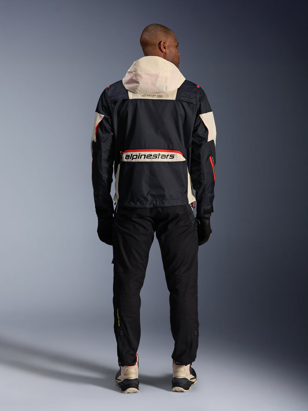 Vista posteriore di un motociclista che indossa la giacca Alpinestars Mogress Air in nero, mandorla e arancione acceso, caratterizzata da un cappuccio color crema, grande marchio Alpinestars sulla tasca posteriore e dettagli delle cerniere arancioni, abbinata a pantaloni neri e stivali color crema per un look da moto urbano.