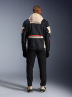 Vista posteriore di un motociclista che indossa la giacca Alpinestars Mogress Air in nero, mandorla e arancione acceso, caratterizzata da un cappuccio color crema, grande marchio Alpinestars sulla tasca posteriore e dettagli delle cerniere arancioni, abbinata a pantaloni neri e stivali color crema per un look da moto urbano.