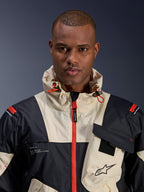 Modello maschile che indossa la giacca Alpinestars Mogress Air, giacca da moto urban, colori Black Almond e Hot Orange, vista ravvicinata che mostra il design color-block, la trama ripstop, il collo con cappuccio con cursori arancioni e i dettagli della cerniera frontale.