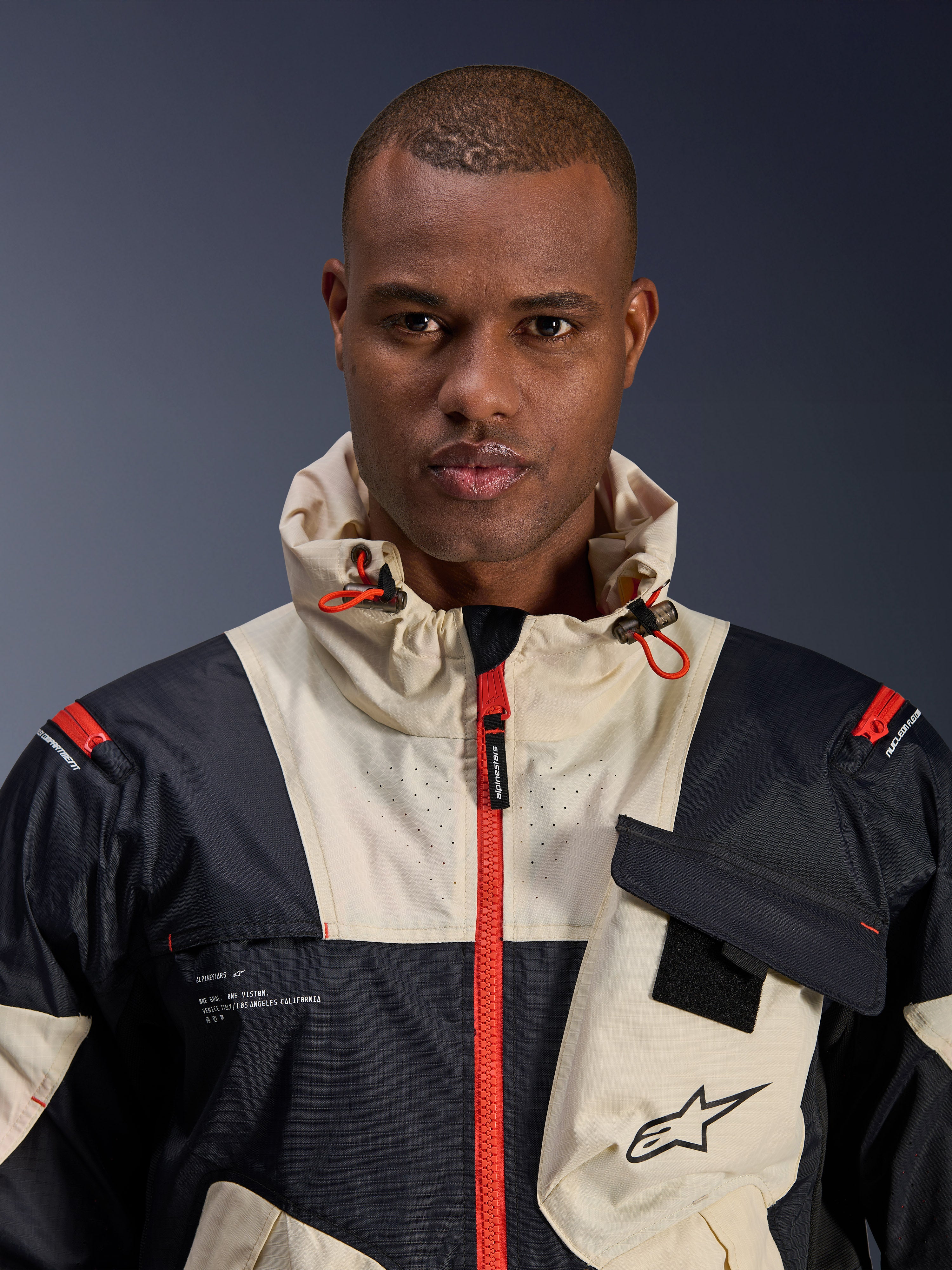 Modello maschile che indossa la giacca Alpinestars Mogress Air, giacca da moto urban, colori Black Almond e Hot Orange, vista ravvicinata che mostra il design color-block, la trama ripstop, il collo con cappuccio con cursori arancioni e i dettagli della cerniera frontale.