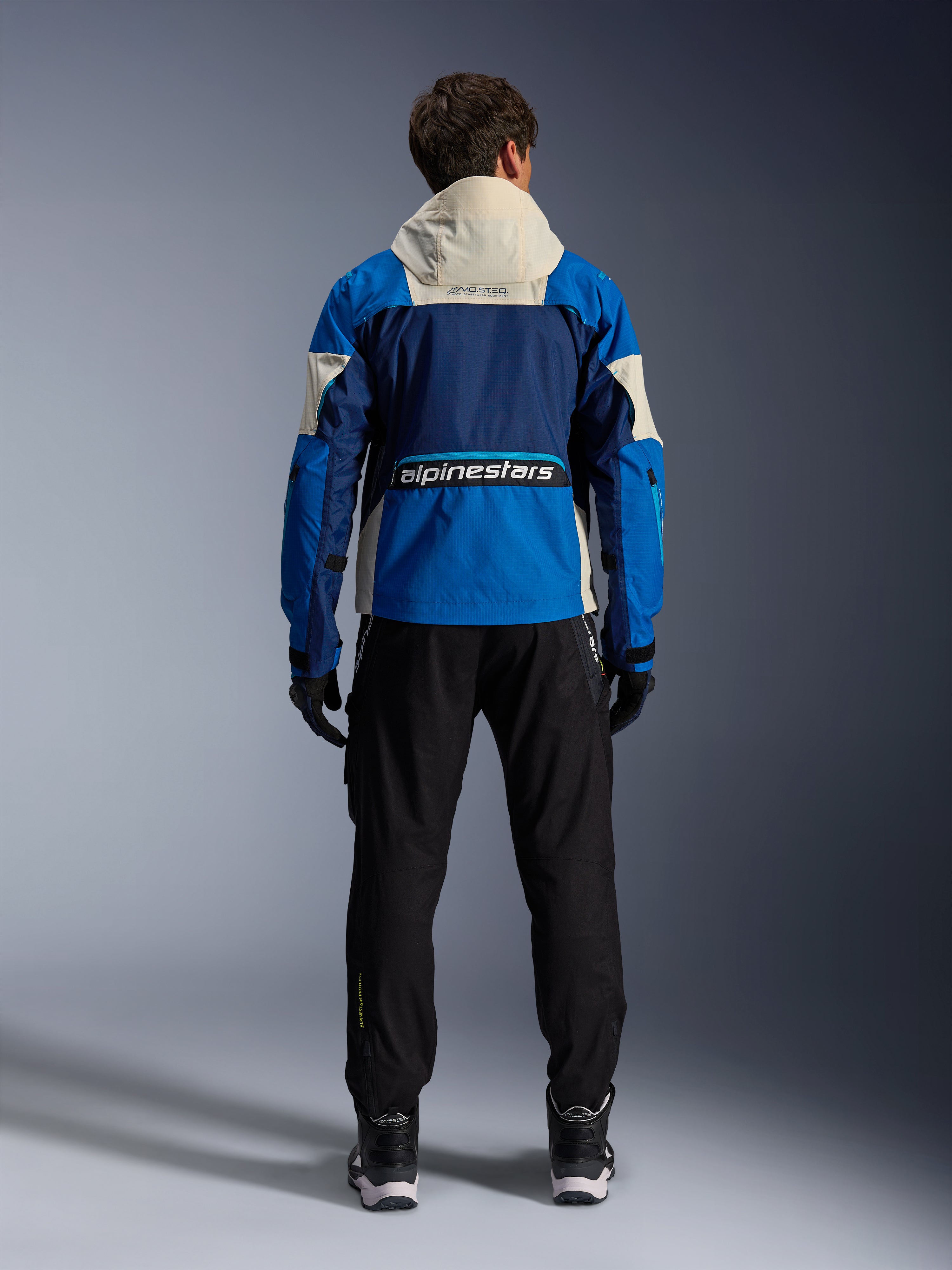 Giacca Alpinestars Mogress Air in Blue Almond, vista posteriore di un motociclista che indossa la giacca da moto, pannelli blu navy e azzurro vivace con cappuccio bianco sporco, logo Alpinestars sulla parte inferiore della schiena, stile urban contemporaneo