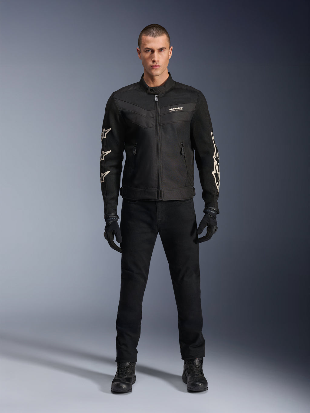 Modello maschile che indossa la giacca Alpinestars T-Dyno Air, nera, giacca da moto leggera in tessuto con mesh 3D traspirante, loghi a stella bianchi sulle maniche, abbinata a pantaloni, guanti e stivali neri, vista frontale in piedi