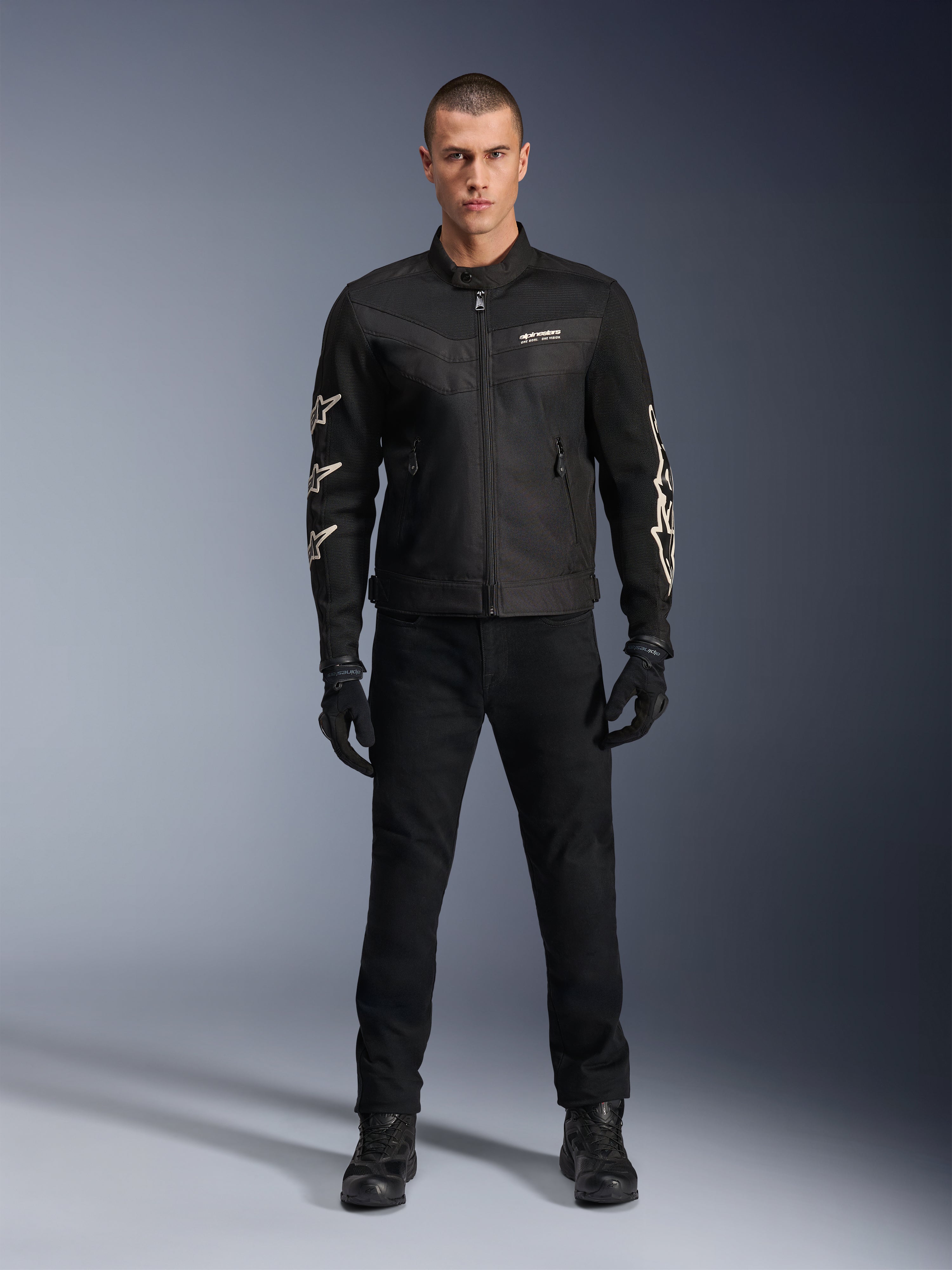 Modello maschile che indossa la giacca Alpinestars T-Dyno Air, nera, giacca da moto leggera in tessuto con mesh 3D traspirante, loghi a stella bianchi sulle maniche, abbinata a pantaloni, guanti e stivali neri, vista frontale in piedi