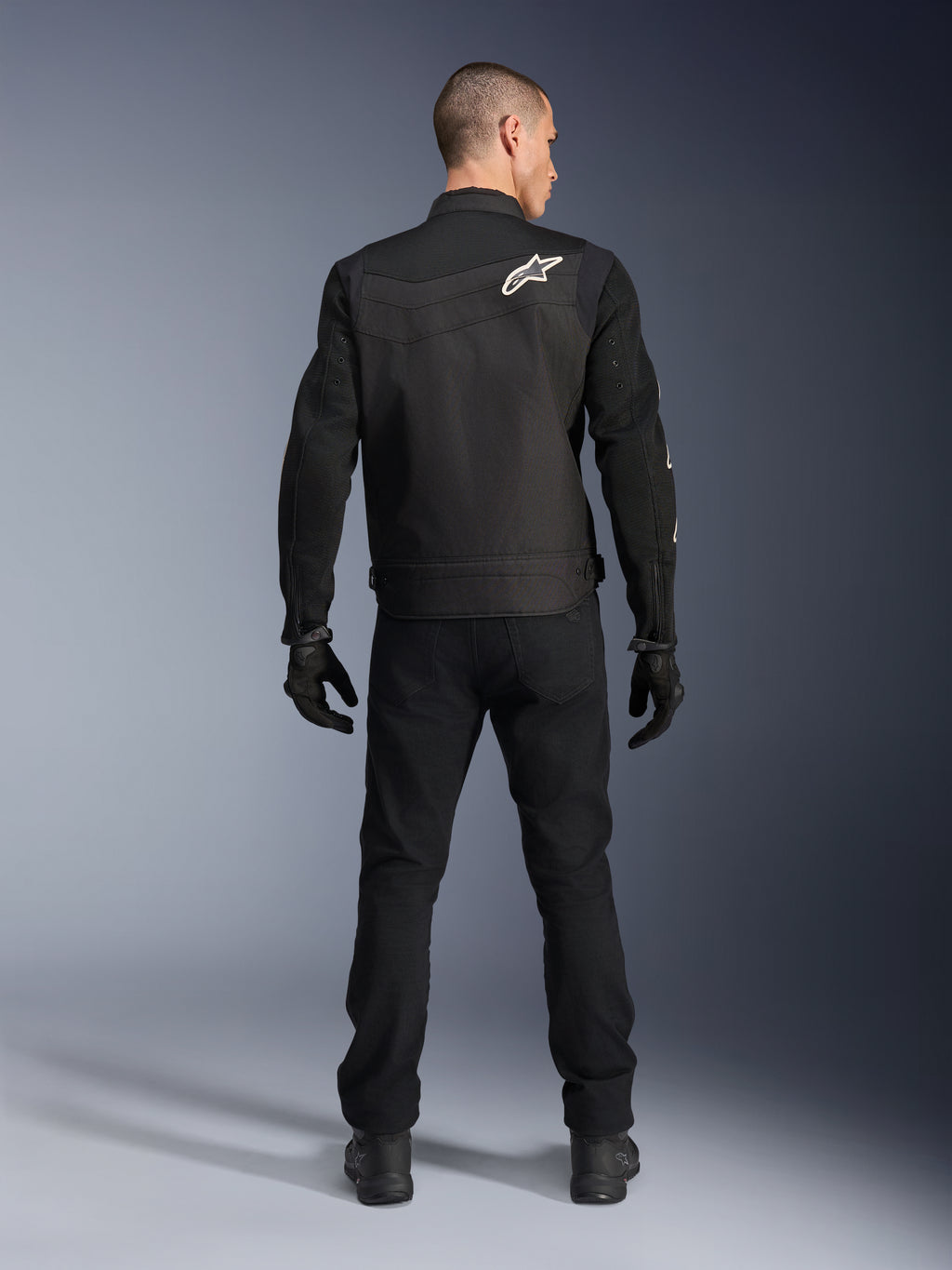 Giacca Alpinestars T-Dyno Air, nera, vista posteriore su modello, giacca da moto in tessuto con pannelli in mesh 3D e logo a stella bianco sulla spalla, indossata con pantaloni e guanti neri, outfit da guida urbana moderno