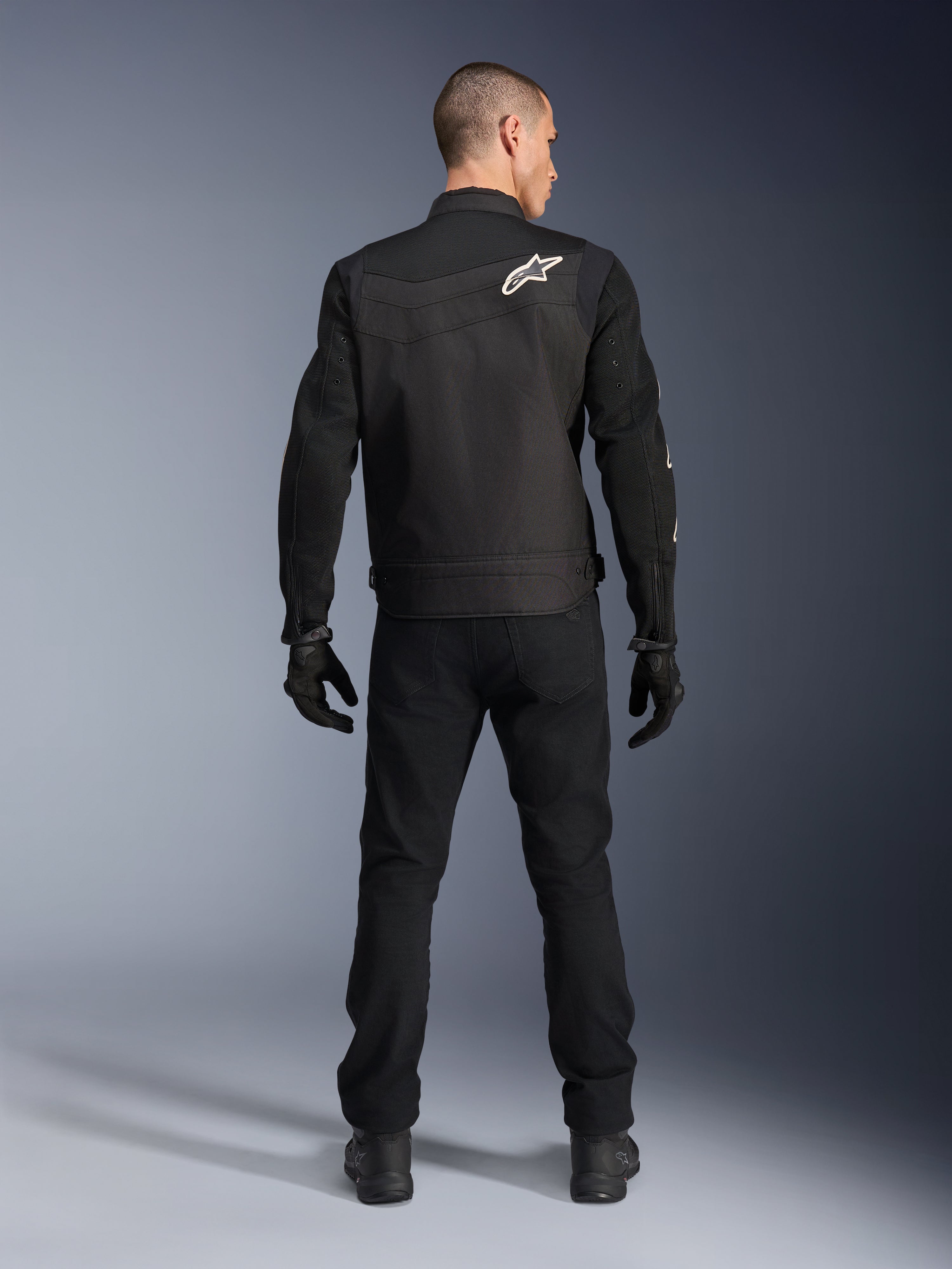 Giacca Alpinestars T-Dyno Air, nera, vista posteriore su modello, giacca da moto in tessuto con pannelli in mesh 3D e logo a stella bianco sulla spalla, indossata con pantaloni e guanti neri, outfit da guida urbana moderno