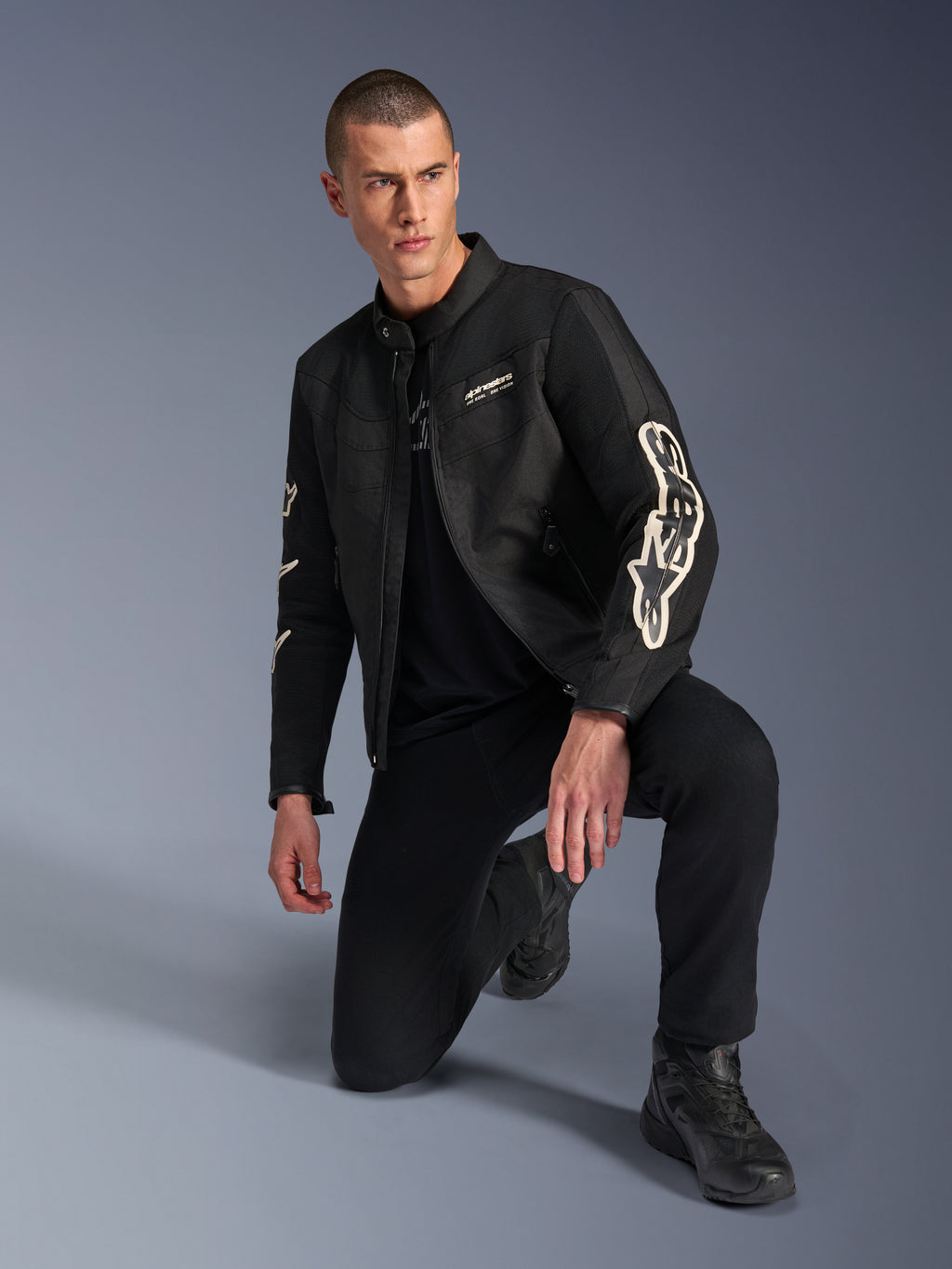 Modello che indossa la giacca Alpinestars T-Dyno Air, giacca da moto in tessuto nero con grande logo bianco sulle maniche, abbinata a pantaloni neri e stivali da moto, design moderno di ispirazione vintage con pannelli in rete 3D