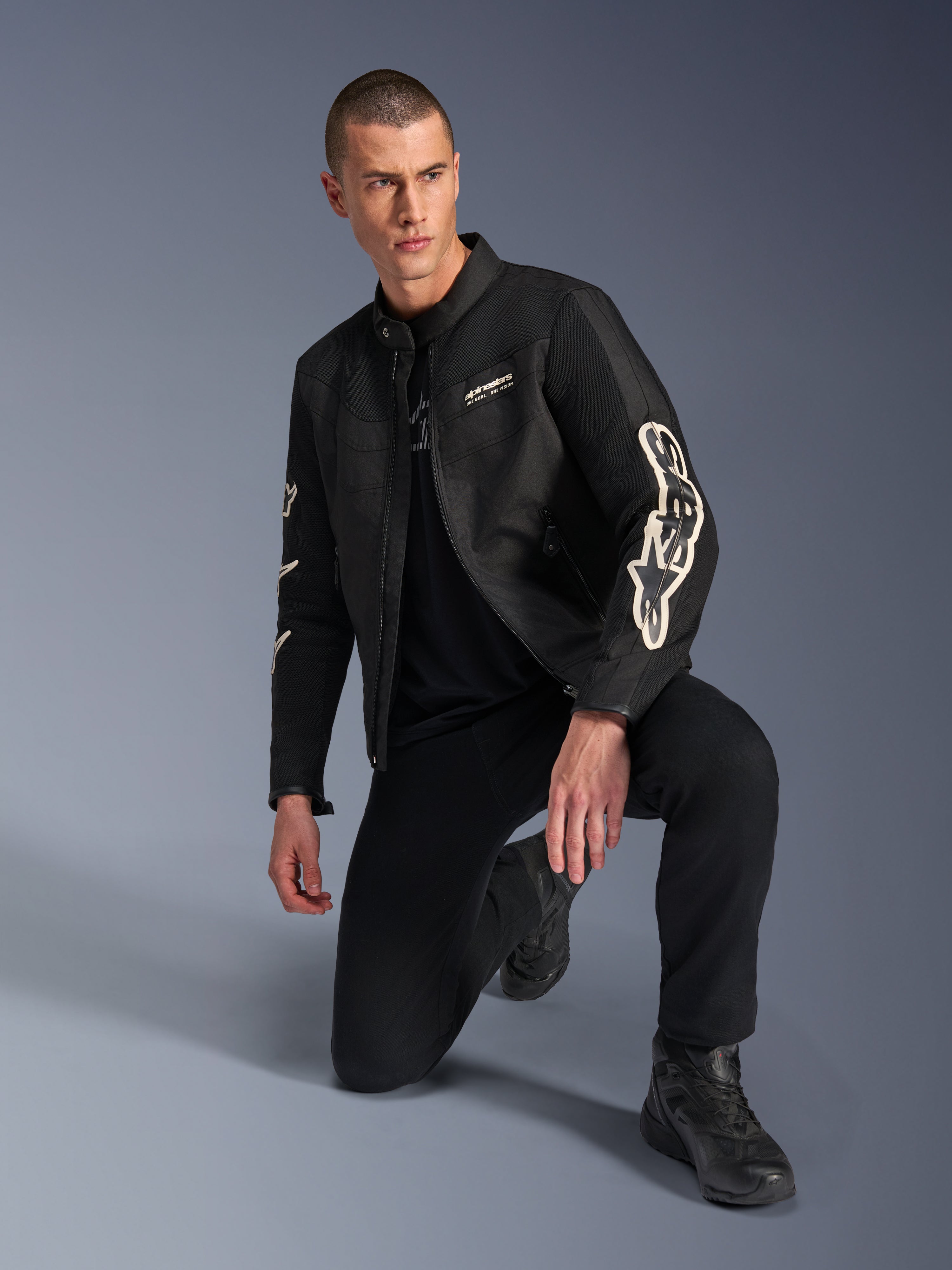Modello che indossa la giacca Alpinestars T-Dyno Air, giacca da moto in tessuto nero con grande logo bianco sulle maniche, abbinata a pantaloni neri e stivali da moto, design moderno di ispirazione vintage con pannelli in rete 3D