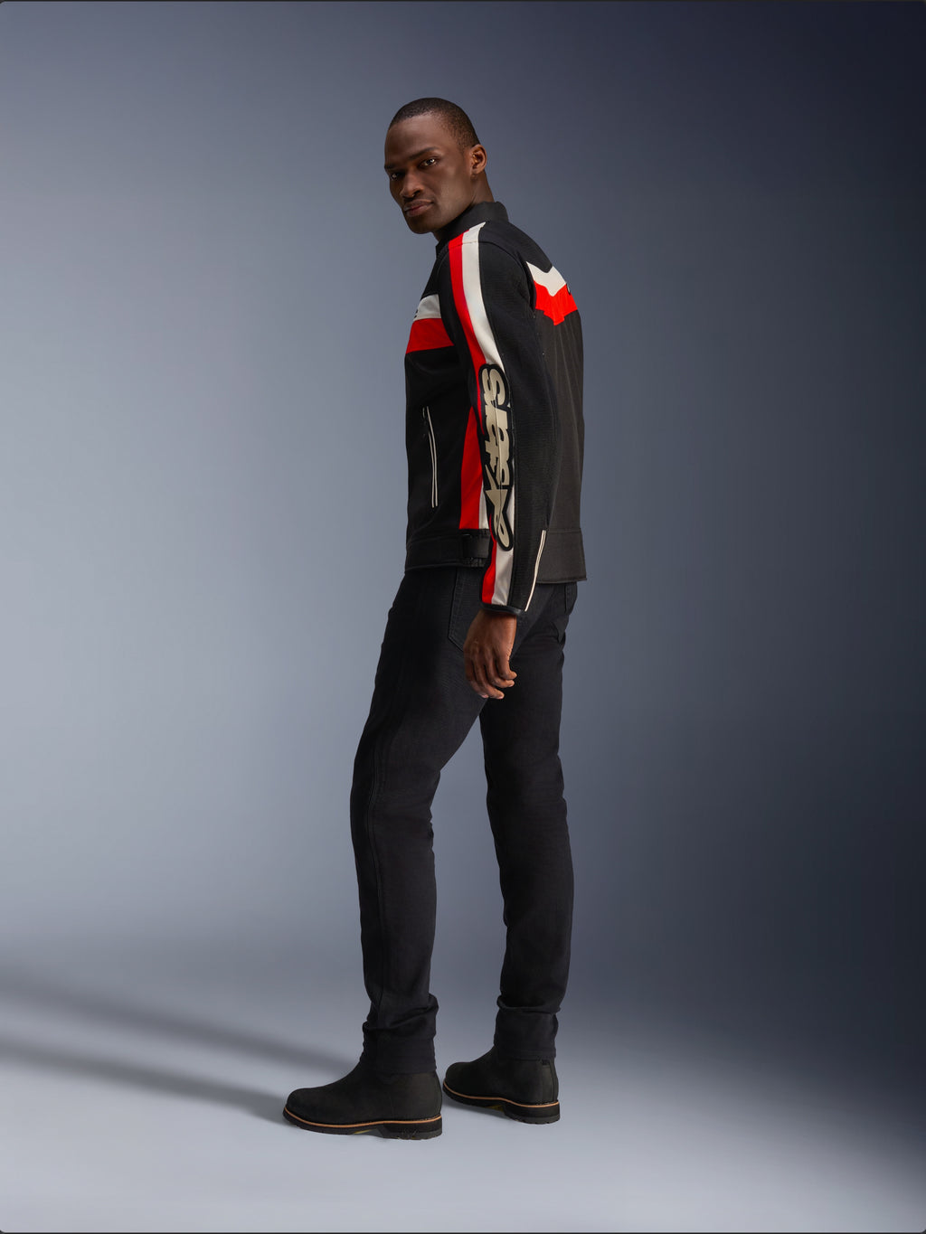 Un uomo indossa la giacca Alpinestars T-Dyno Air in nero, rosso acceso ed ecru, giacca da moto in tessuto d'ispirazione vintage con pannelli in mesh, logo vistoso sulla manica, abbinata a pantaloni e stivali neri per un moderno look da motociclista