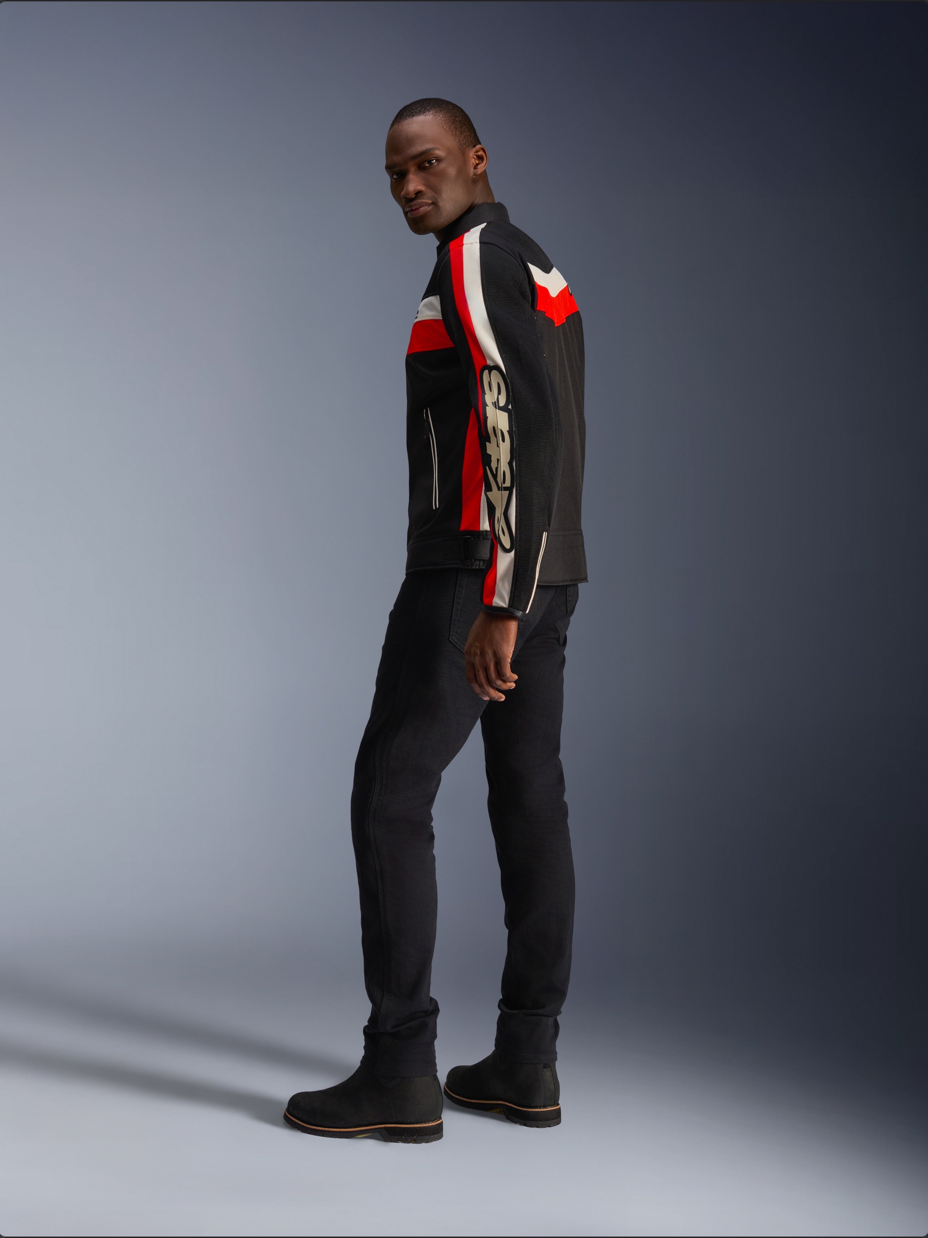 Un uomo indossa la giacca Alpinestars T-Dyno Air in nero, rosso acceso ed ecru, giacca da moto in tessuto d'ispirazione vintage con pannelli in mesh, logo vistoso sulla manica, abbinata a pantaloni e stivali neri per un moderno look da motociclista