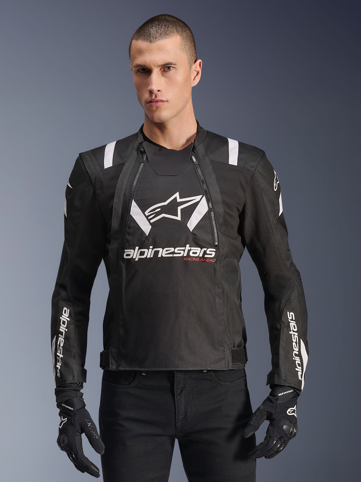 Alpinestars T-Stunt Air Jacket, giacca da moto bianca e nera, indossata da un uomo con guanti e pantaloni neri, con branding bianco evidente su petto e maniche e accenti rossi 'Racing Ahead', design sportivo con strisce bianche sulle spalle e pannelli in rete traspirante.