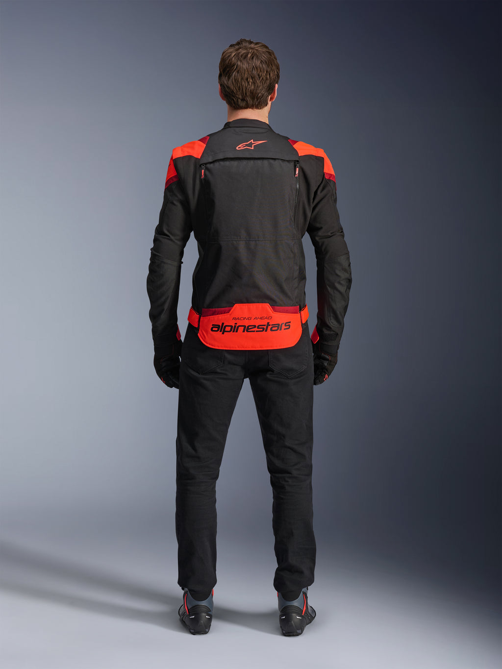 Giacca Alpinestars T-Stunt Air, nero con accenti rosso fluo e rosso scuro, vista posteriore su modello, abbinata a pantaloni da moto e stivali neri, con marchio Alpinestars sul pannello posteriore inferiore, design modulare con costruzione in mesh per la ventilazione.