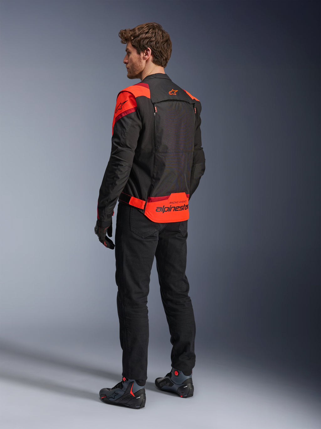 Uomo che indossa la giacca Alpinestars T-Stunt Air vista da dietro, nero, rosso fluo e rosso scuro, giacca da moto in tessuto con pannelli in mesh e design dal taglio sportivo, con vistoso marchio Alpinestars sulla parte inferiore della schiena, abbinata a pantaloni neri e stivali da guida