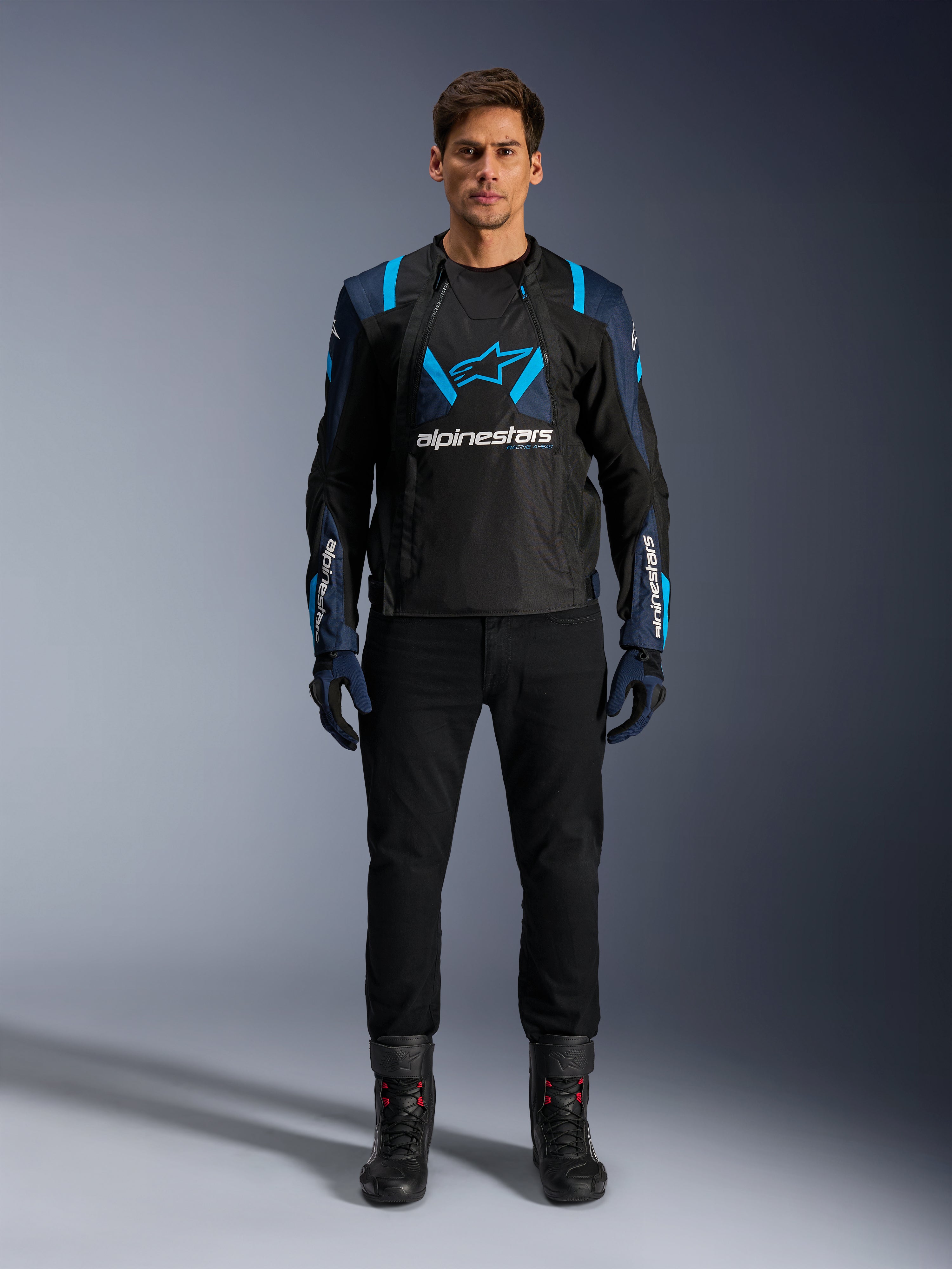 Giacca Alpinestars T-Stunt Air, giacca da moto, nera con accenti blu scuro e azzurro brillante, con un grande logo bianco sul petto, costruzione in mesh a taglio sportivo per il flusso d'aria, indossata con pantaloni da moto e stivali neri per un'estetica road riding elegante.