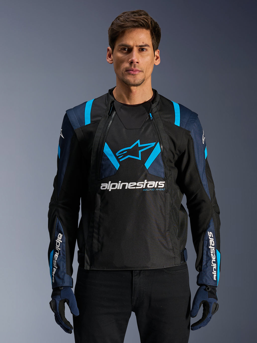 Alpinestars T-Stunt Air Jacket, giacca da moto in rete, nero e blu, indossata da un modello con pantaloni e guanti neri, design modulare 2-in-1 con pannelli in mesh tecnico traspirante, logo a stella sul petto e marchio Alpinestars sulle maniche.