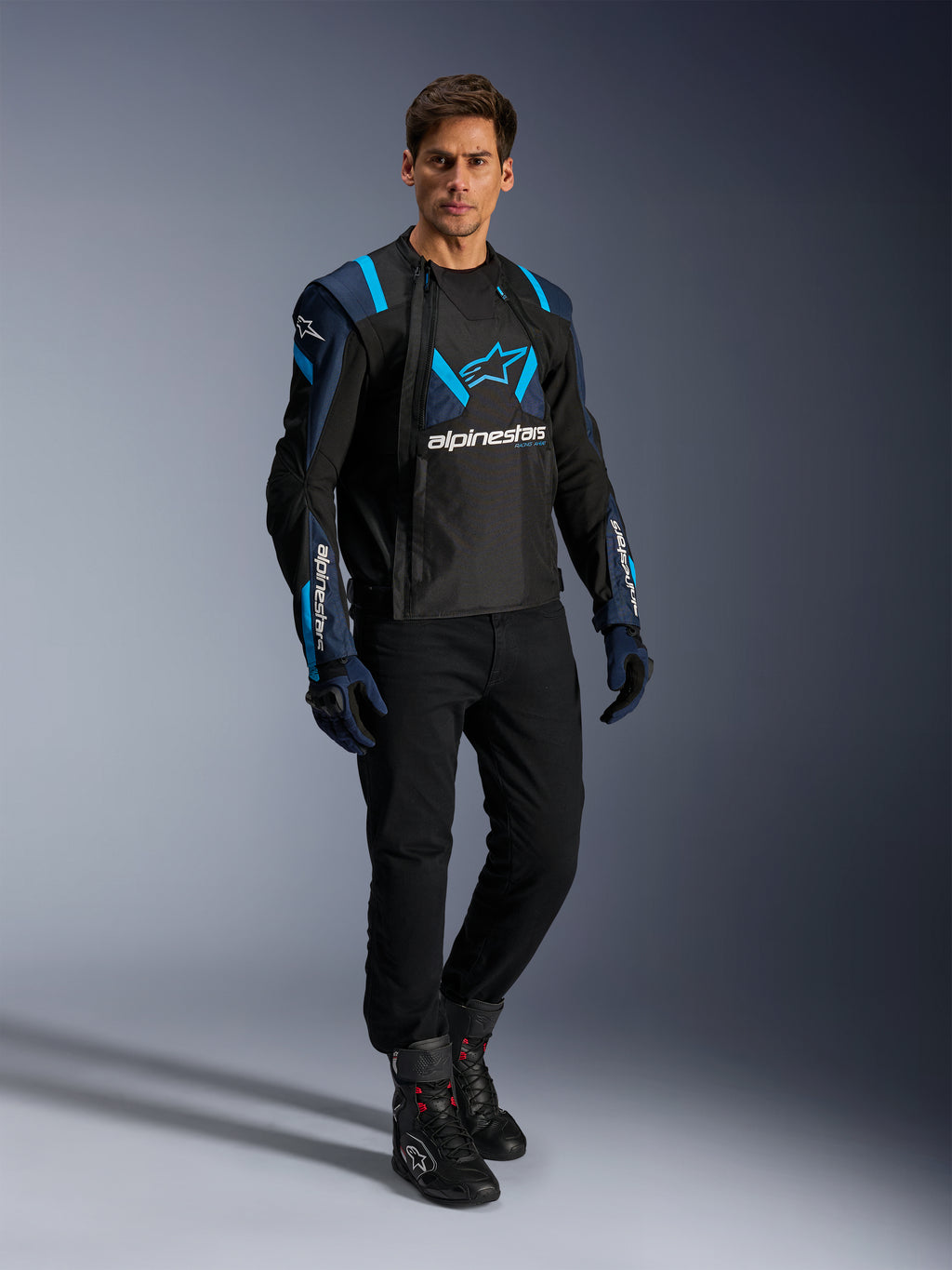 Modello che indossa la giacca Alpinestars T-Stunt Air, nero, blu scuro e azzurro, con evidenti pannelli in mesh per il flusso d'aria, abbinata a pantaloni neri e guanti da moto, giacca da moto dal taglio sportivo per la guida su strada estiva.