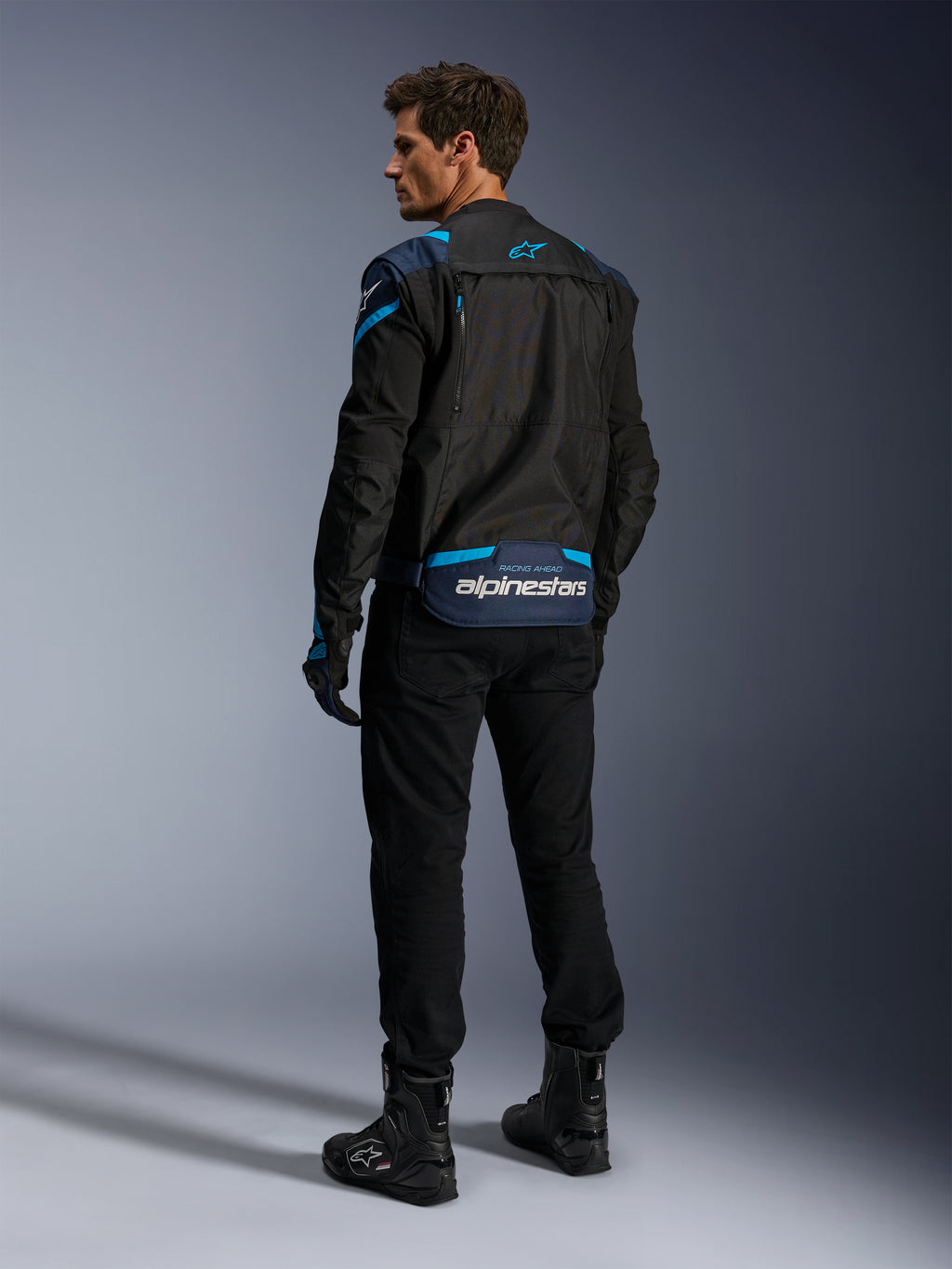 Alpinestars Giacca T-Stunt Air, giacca da moto, nero con accenti blu scuro e azzurro, vista posteriore che mostra i pannelli modulari in rete e il marchio sulla parte inferiore della schiena, indossata da un modello con pantaloni da guida e stivali neri.