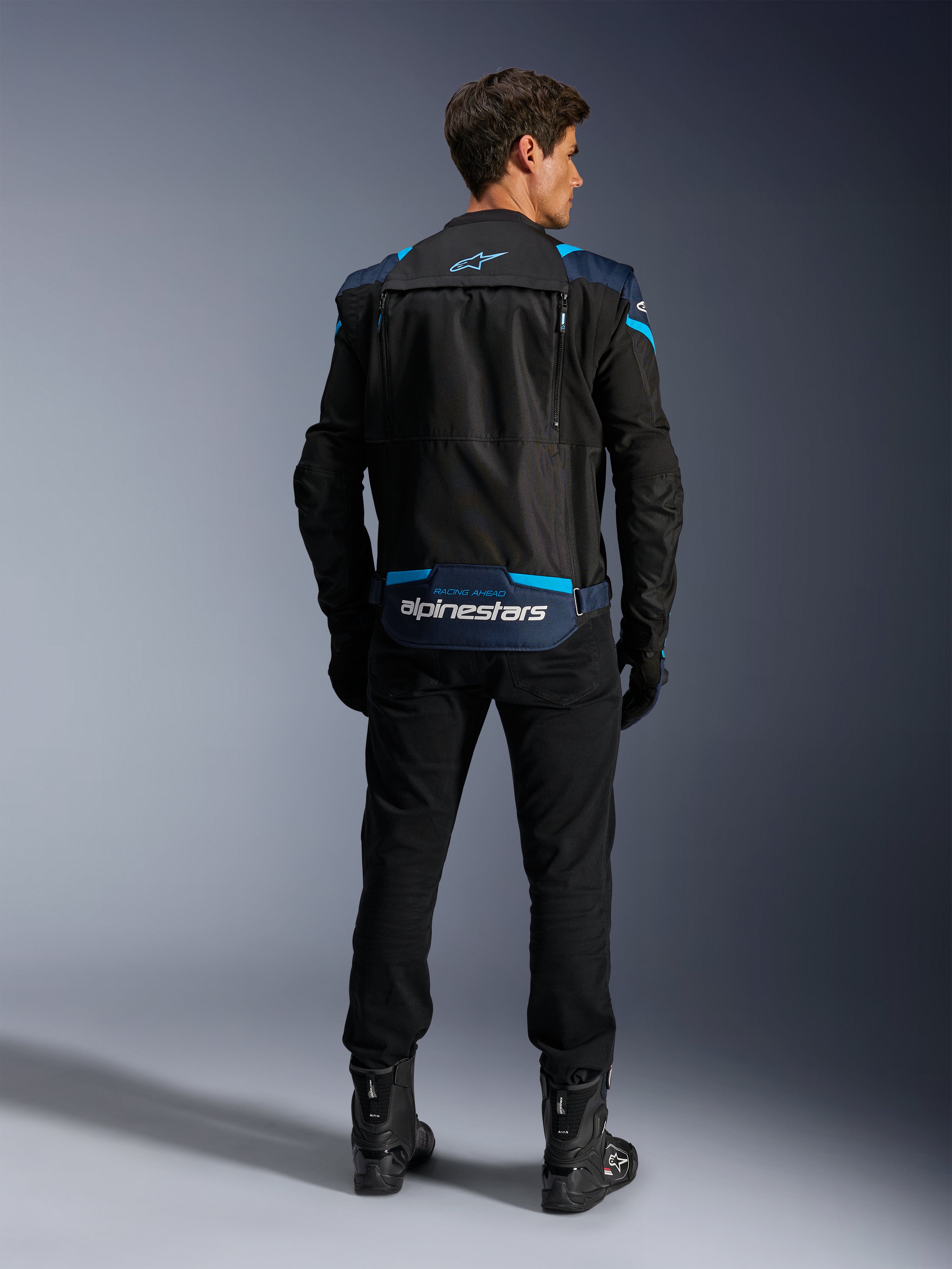 Alpinestars T-Stunt Air Jacket, giacca da moto modulare in tessuto, nero, blu scuro e blu brillante, vista posteriore che mostra lo scomparto inferiore per le maniche con logo Alpinestars bianco, indossata con pantaloni e stivali neri.
