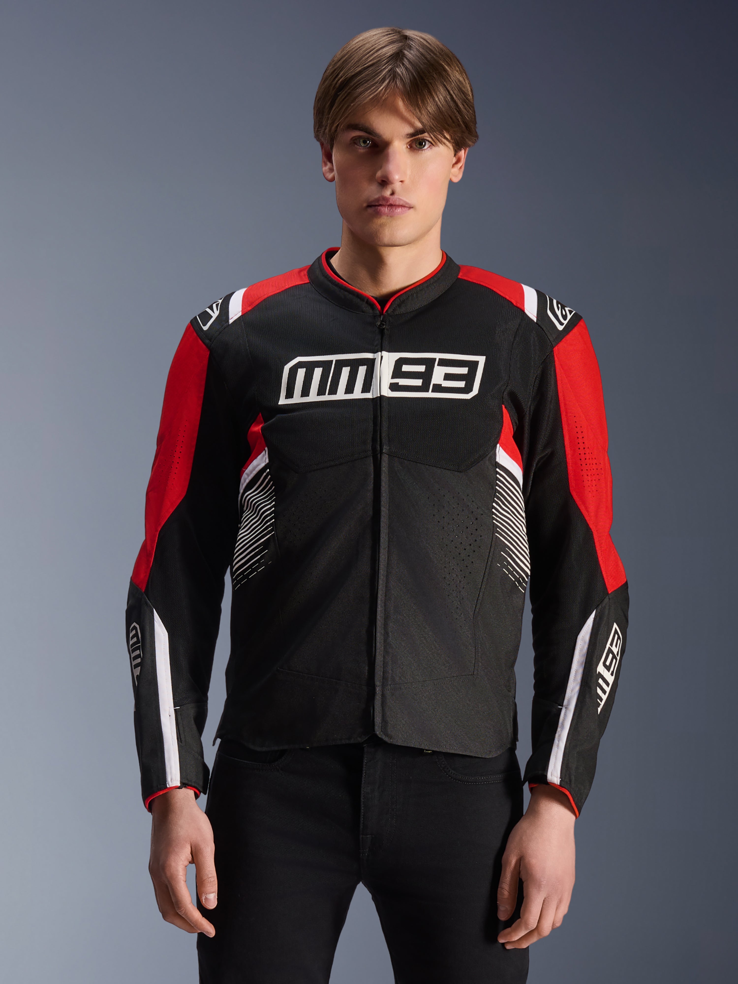 Giacca MM93 T-SPS Air V2, giacca da moto in tessuto in nero, rosso acceso e bianco, con marchio MM93 di Marc Márquez su petto e maniche, pannelli in mesh per la ventilazione, design dal taglio sportivo, indossata da un modello con pantaloni neri