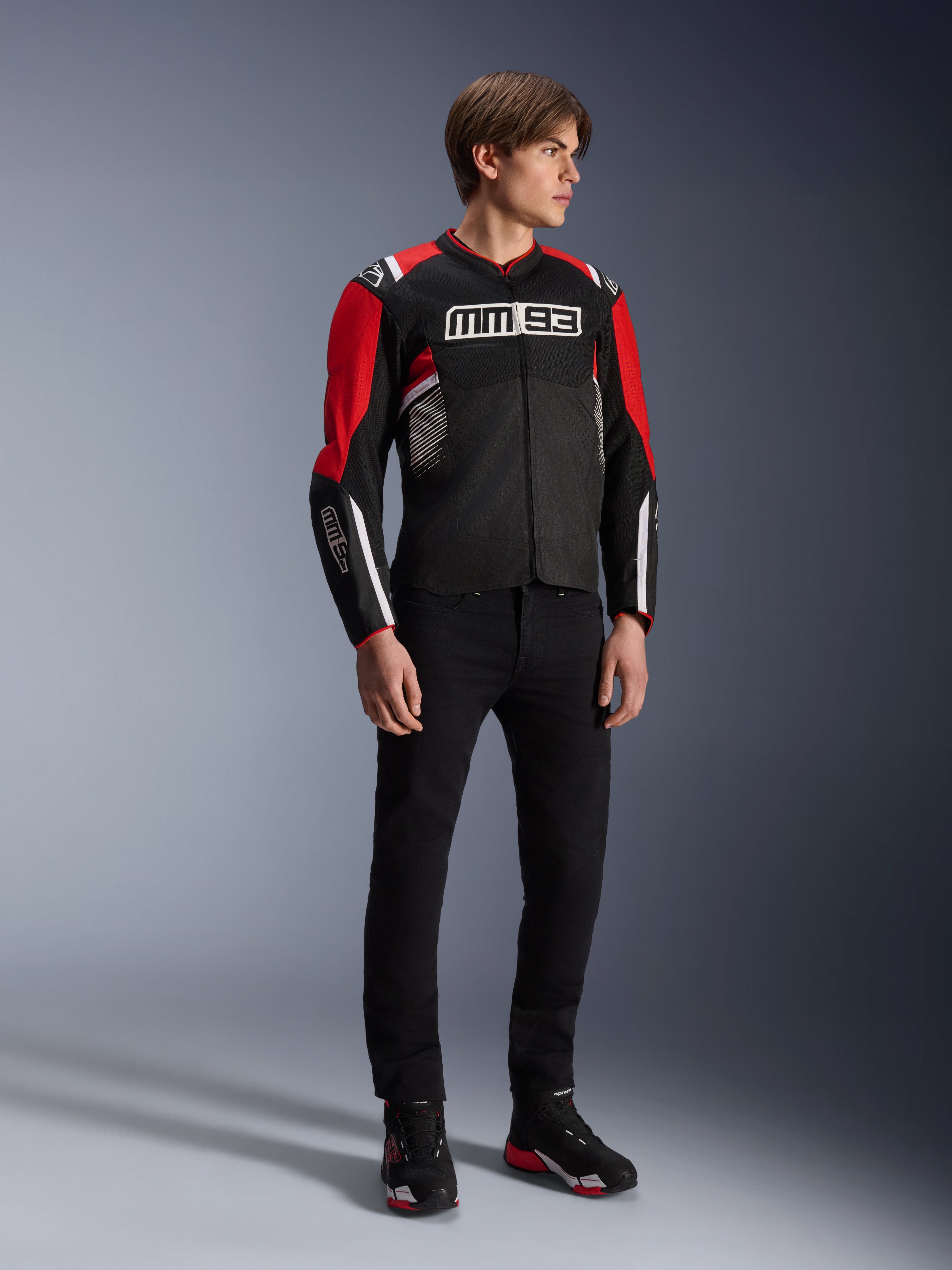 Modello che indossa la giacca Alpinestars MM93 T-SPS Air v2 in nero, rosso acceso e bianco, con marchio MM93 su petto e maniche, abbinata a pantaloni neri e stivali da moto, stile urbano sportivo con pannelli in rete per il flusso d'aria