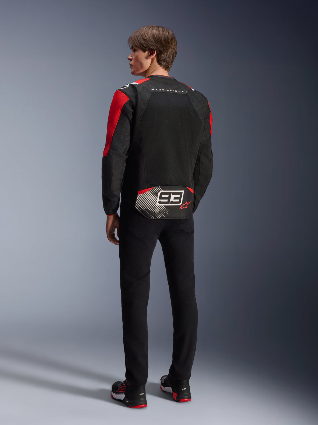 Modello di spalle che indossa la giacca Alpinestars MM93 T-SPS Air V2, nera con dettagli rosso brillante e bianchi, con scritta 'MARC MARQUEZ' e grande logo '93' sulla parte posteriore, struttura in tessuto mesh per la ventilazione, abbinata a pantaloni neri e scarpe da moto coordinate nere e rosse.