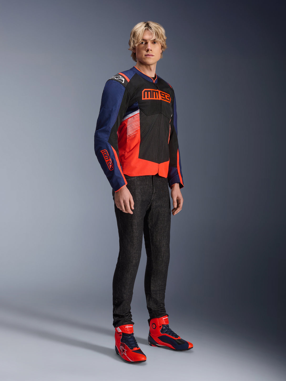 Modello che indossa la giacca Alpinestars MM93 T-SPS Air V2 in blu navy, nero e rosso fluo, con il vistoso marchio MM93 sul petto e sulle maniche, abbinata a jeans scuri e stivali da moto coordinati rossi e blu, abbigliamento sportivo per guida urbana