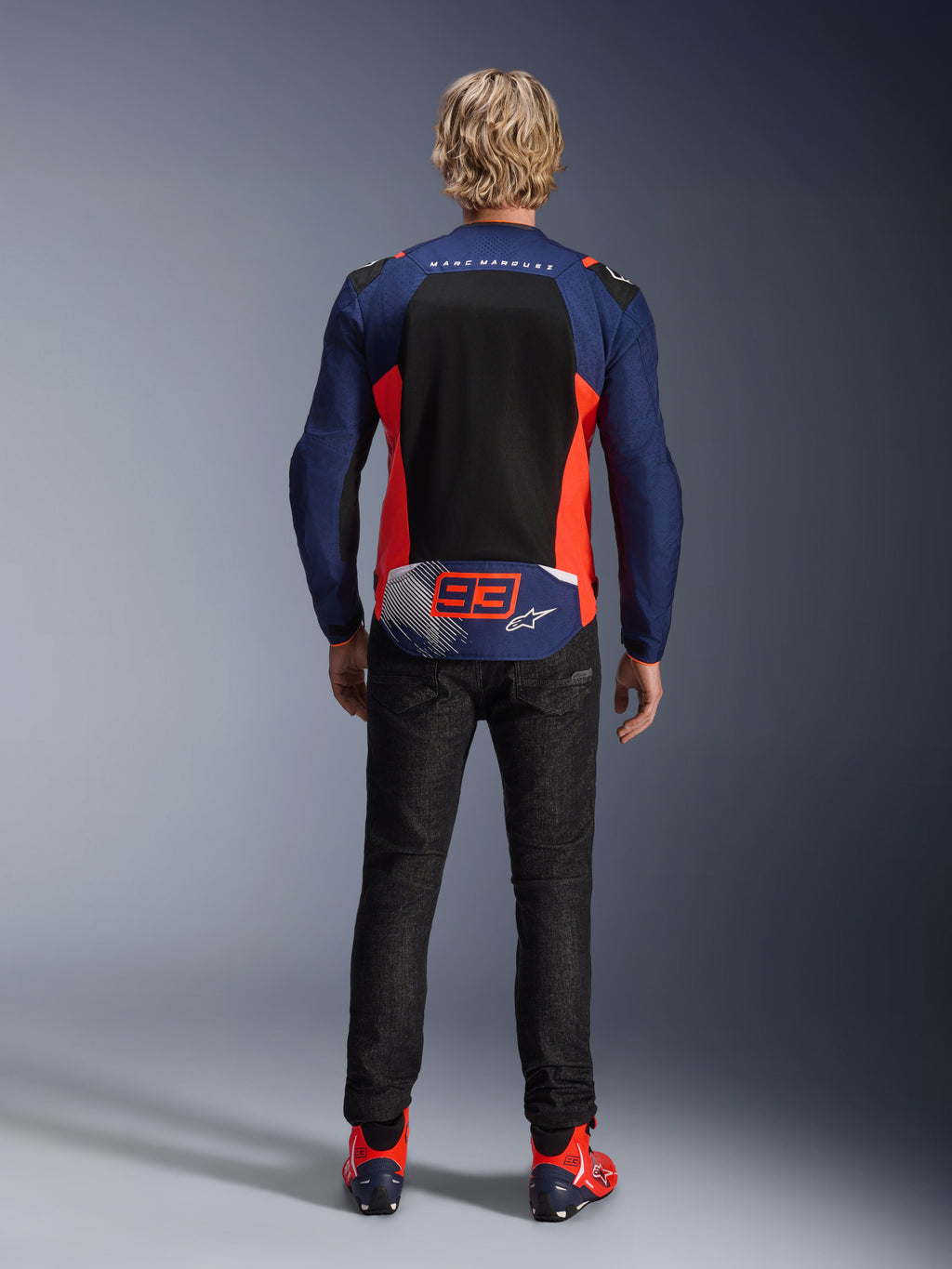 Vista posteriore di un modello che indossa la giacca MM93 T-SPS Air V2 in blu navy con accenti rosso fluo e nero, caratterizzata da un ampio pannello posteriore in rete e il marchio '93' di Marc Marquez, abbinata a pantaloni in denim scuro e stivali da moto coordinati.