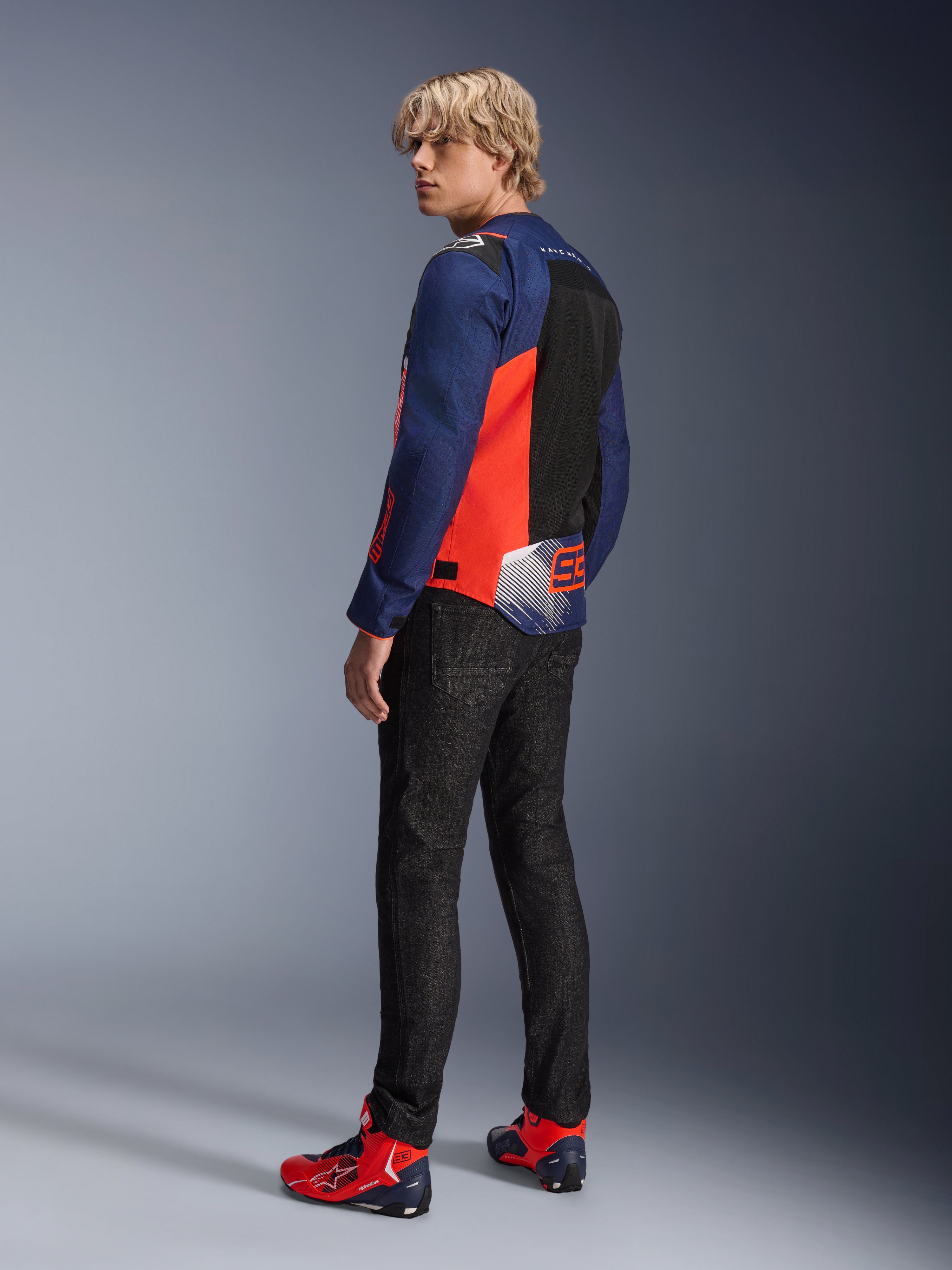 Giacca Alpinestars MM93 T-SPS Air V2, blu navy e rosso fluo, indossata da un modello vista posteriore con jeans da moto neri e scarpe coordinate rosse e blu, dotata di pannelli in mesh e logo Marc Márquez 93.