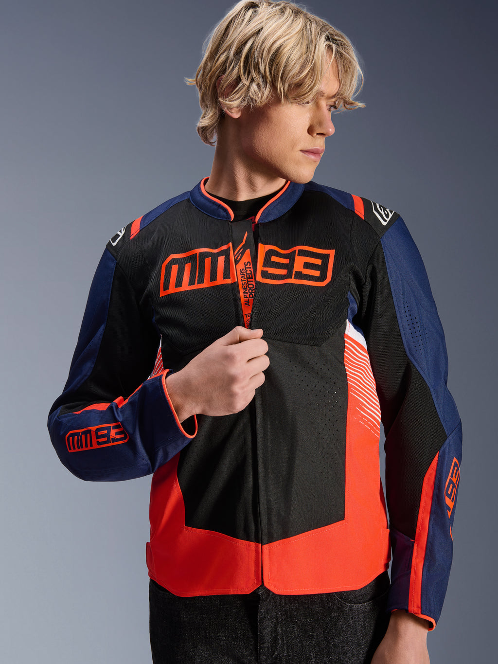 Giacca Alpinestars MM93 T-SPS Air V2, giacca da moto in tessuto blu navy, nero e rosso fluorescente, indossata da un modello mentre regola la zip, con marchio Marc Márquez MM93 su petto e maniche e pannelli in mesh per ventilazione estiva.