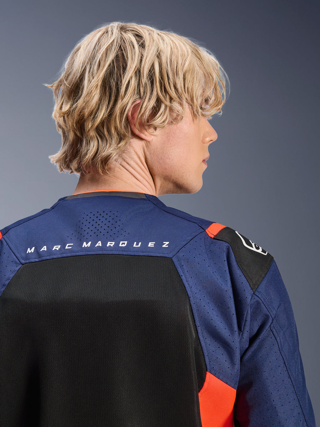 Giacca Alpinestars MM93 T-SPS Air V2, blu navy e nero con accenti rosso fluo, primo piano della parte superiore della schiena con marchio 'MARC MARQUEZ', pannelli in rete traspirante e costruzione in tessuto traforato, indossata da un modello biondo.