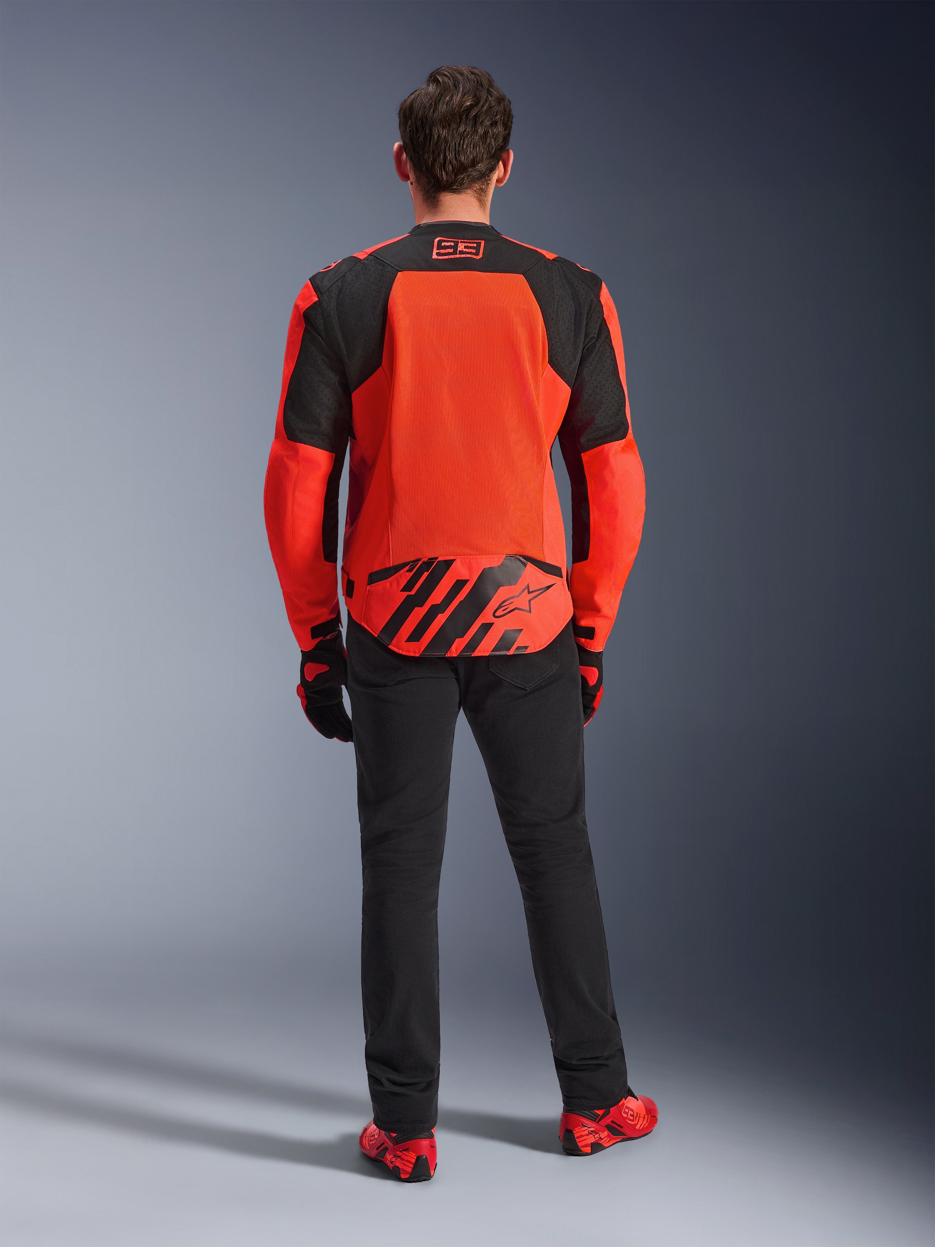 Vista posteriore della giacca MM93 T-SPS Air V2, Rosso Fluo e Nero, giacca da moto in tessuto con ampi pannelli in rete sulla schiena per la ventilazione, indossata con pantaloni neri e calzature rosse, con marchio Marc Marquez 93