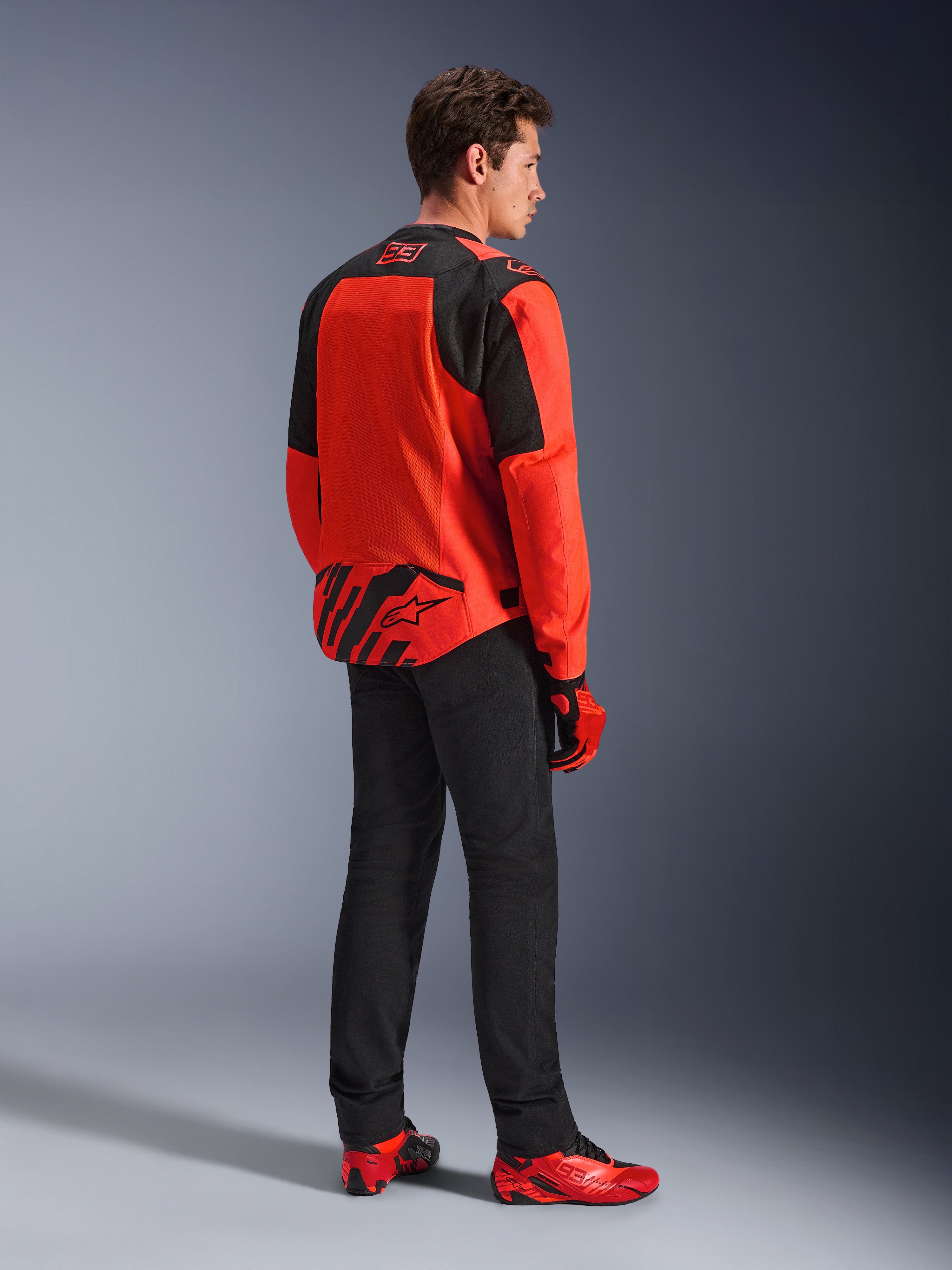 Giacca Alpinestars MM93 T-SPS Air V2 in Rosso Fluo e Nero, vista posteriore di tre quarti su un modello, con marchio Marc Marquez 93 sul colletto e sulla parte inferiore della schiena, abbinata a pantaloni neri e scarpe da moto rosse.