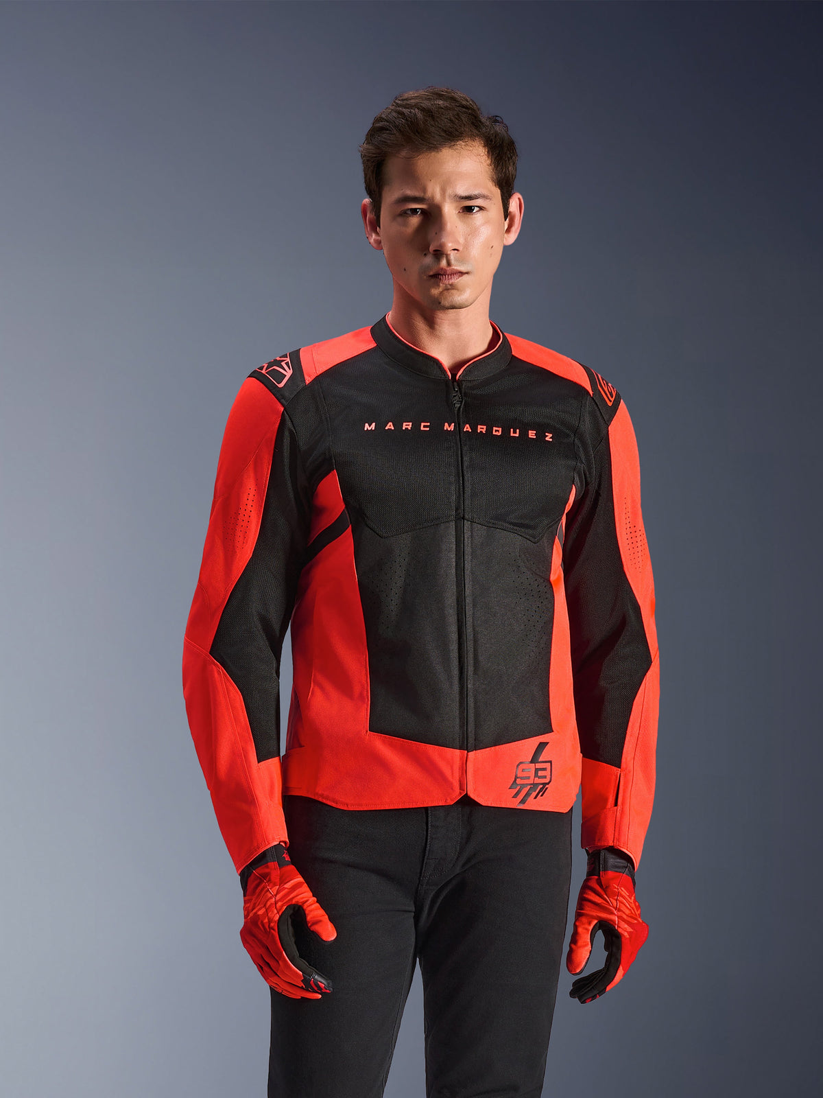 Alpinestars MM93 T-SPS Air V2 Jacket, giacca da moto, rosso fluo e nero, indossata da un modello con guanti rossi coordinati e pantaloni neri, con marchio 'Marc Marquez' sul petto e logo '93', costruzione sportiva in rete