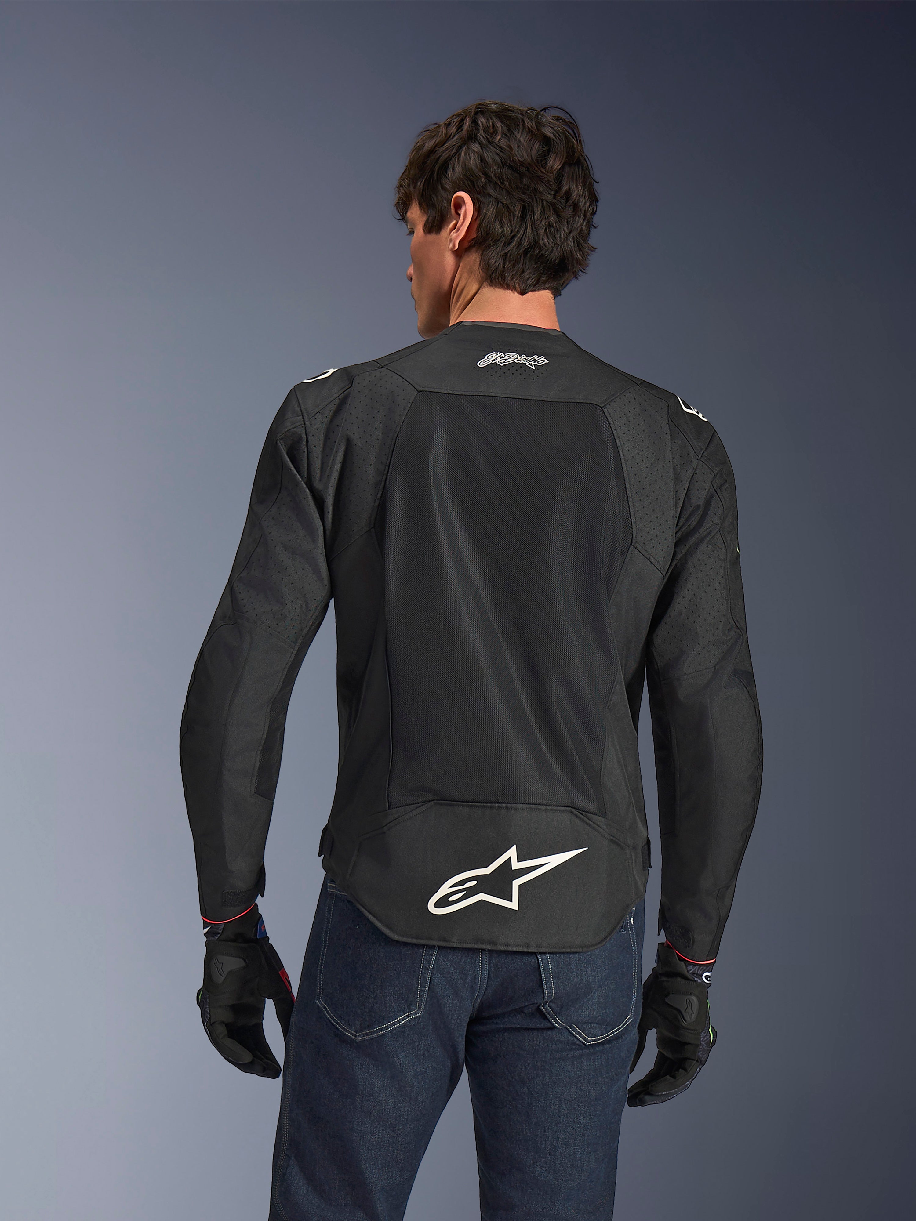 Alpinestars FQ20 T-SPS Air V2 Monster Jacket, nero, vista posteriore con ampi pannelli di ventilazione in rete e logo Alpinestars bianco sulla parte inferiore della schiena, indossata con jeans blu e guanti da moto neri, stile urban sport.