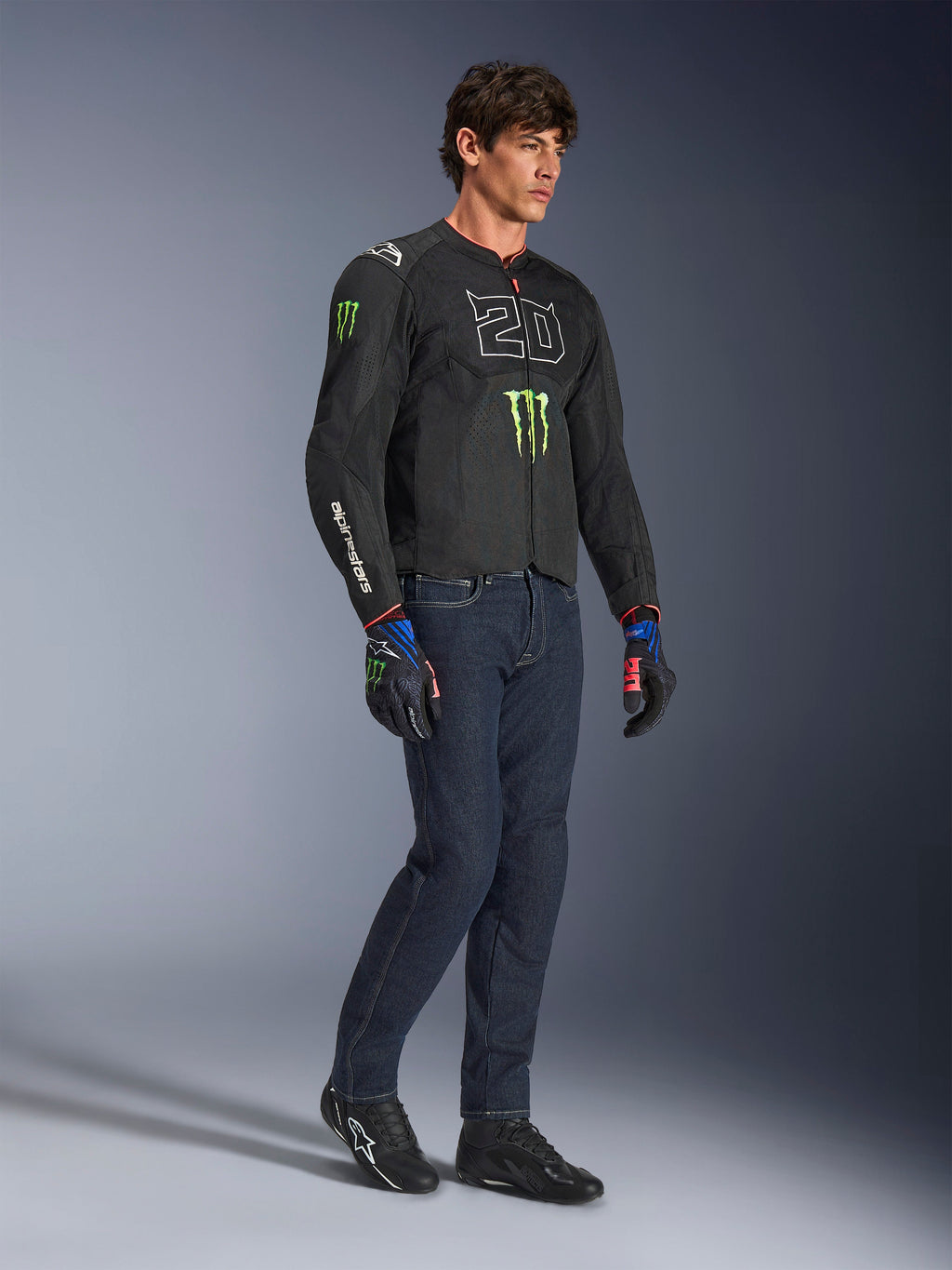 Pilota uomo che indossa la giacca Alpinestars FQ20 T-SPS Air V2 Monster in nero, con il logo '20' e l'artiglio Monster Energy sul petto, abbinata a jeans da moto scuri, scarpe da moto nere e guanti FQ20 coordinati, scatto in studio a figura intera.